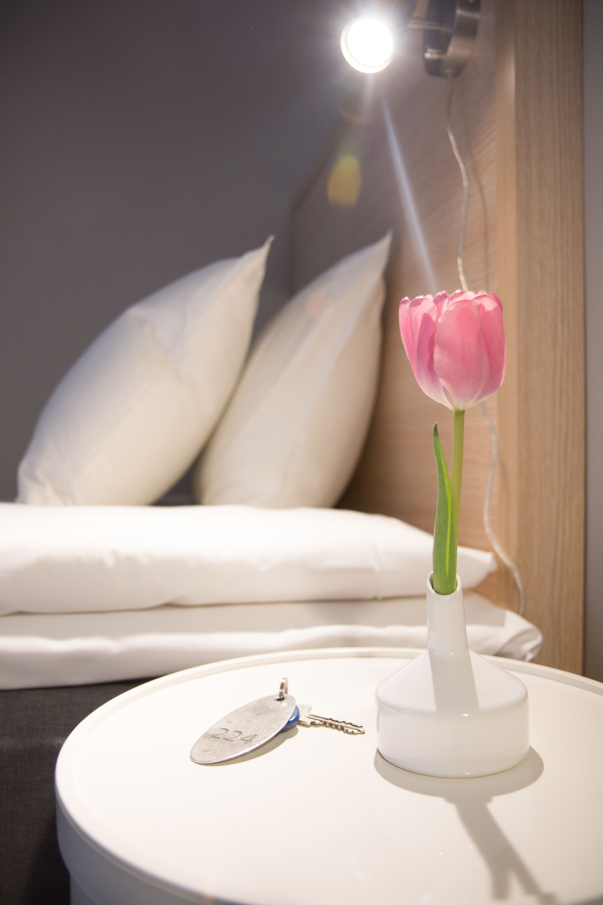Uma mesa de cabeceira branca com uma tulipa rosa num vaso branco, um chaveiro e um abajur com cúpula branca.