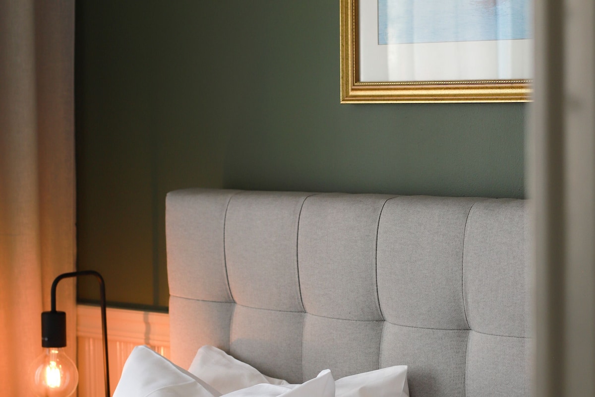 A neatly made bed with white and green pillows, a grey upholstered headboard, and a framed picture on the wall.