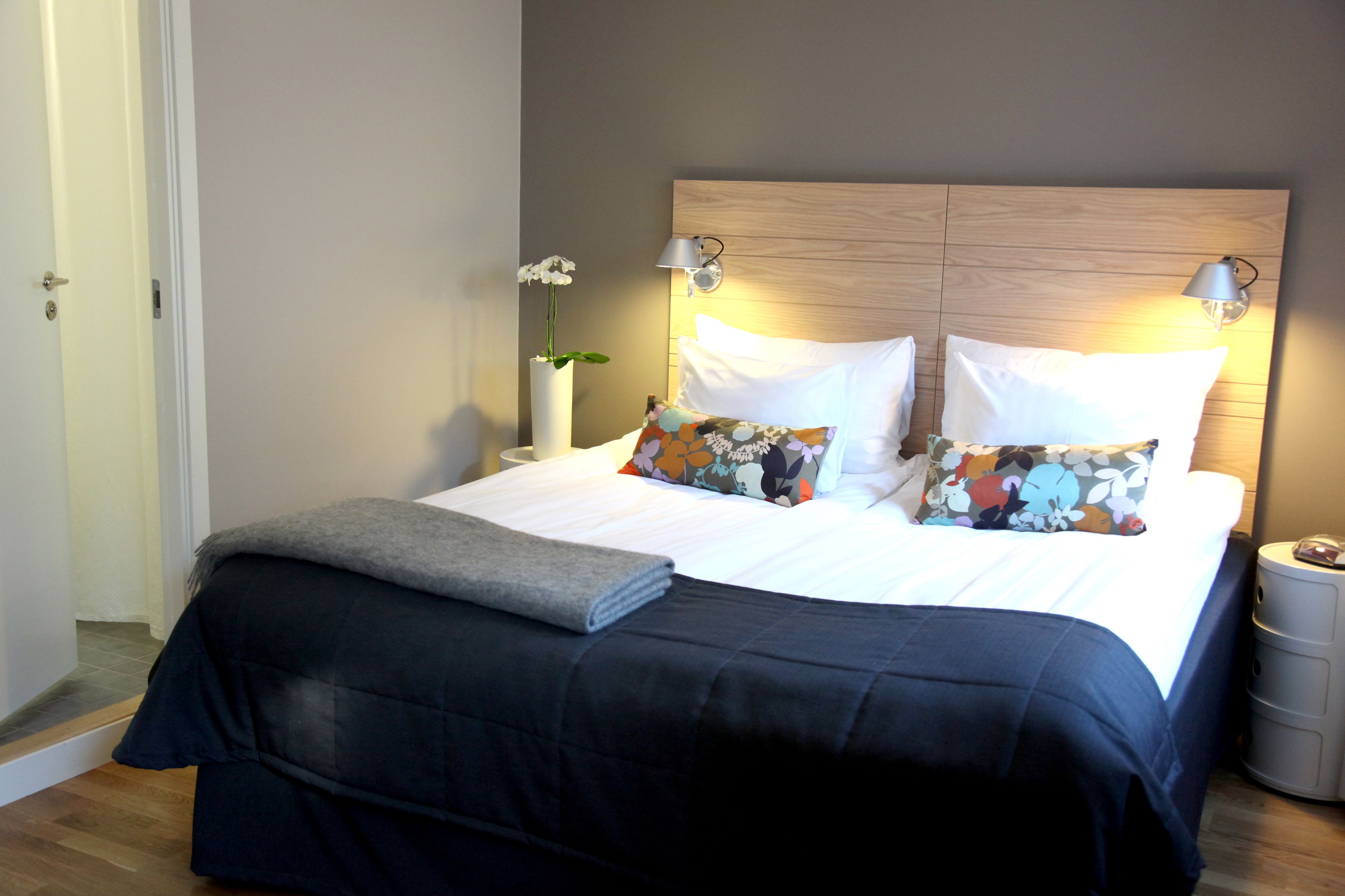 A neatly made bed with a dark blue bedspread, white pillows, and a floral pillow. There is a small table with a lamp and a vase of flowers beside the bed.
