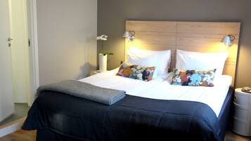 A neatly made bed with a dark blue bedspread, white pillows, and a floral pillow. There is a small table with a lamp and a vase of flowers beside the bed.