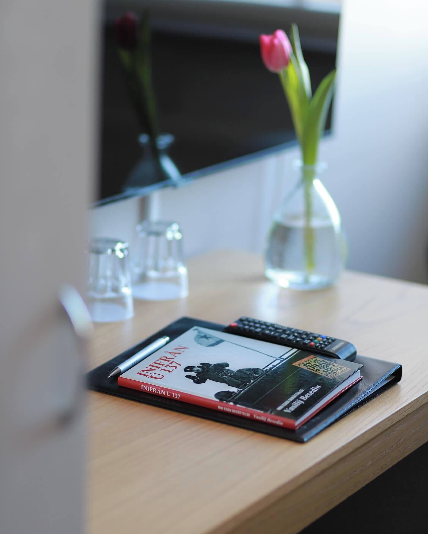 Una tableta, un control remoto y un libro sobre una mesa de madera, con un jarrón de flores al fondo.