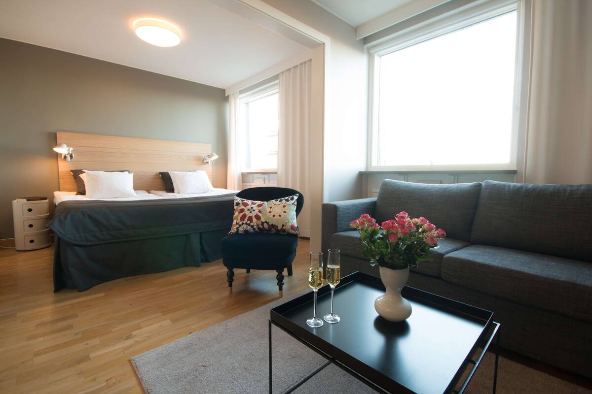 A hotel room with a bed, a chair, a sofa, a coffee table with a vase of flowers, and a tray with glasses.