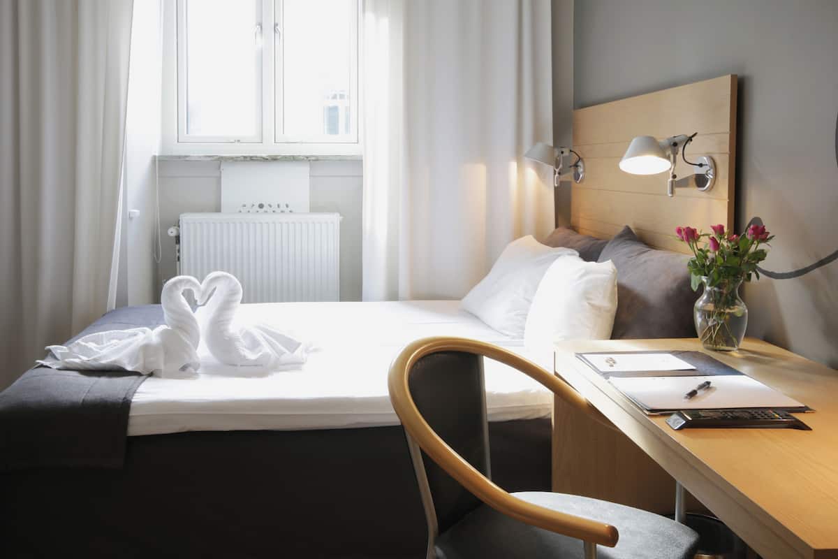 A hotel room with a bed, a desk, a chair, a radiator, a window, and a wall-mounted lamp.
