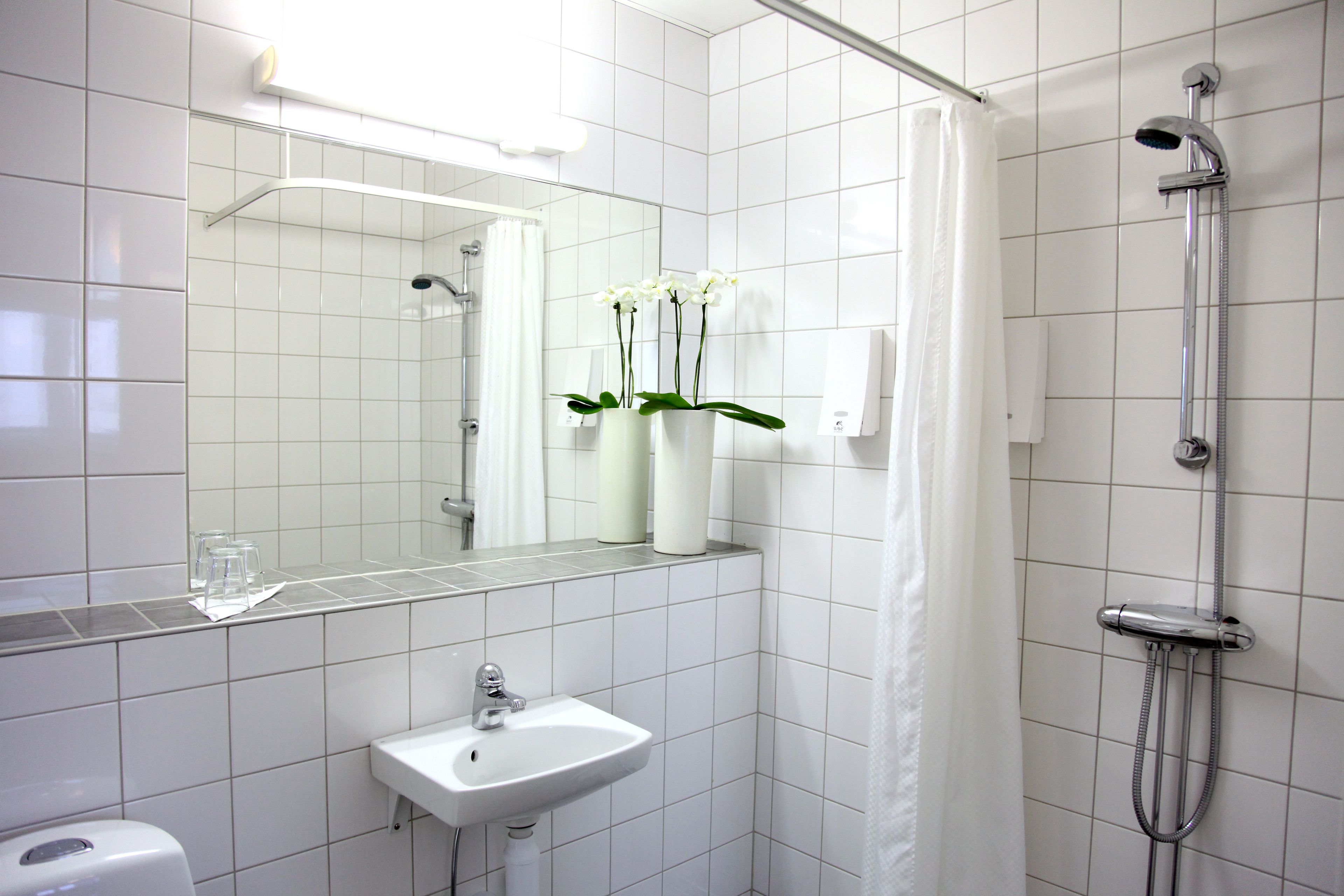 A bathroom with a white sink, mirror, and shower. There are two white vases with orchids on the counter.