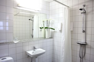 Un baño con lavamanos blanco, espejo y ducha. Hay dos jarrones blancos con orquídeas en la encimera.