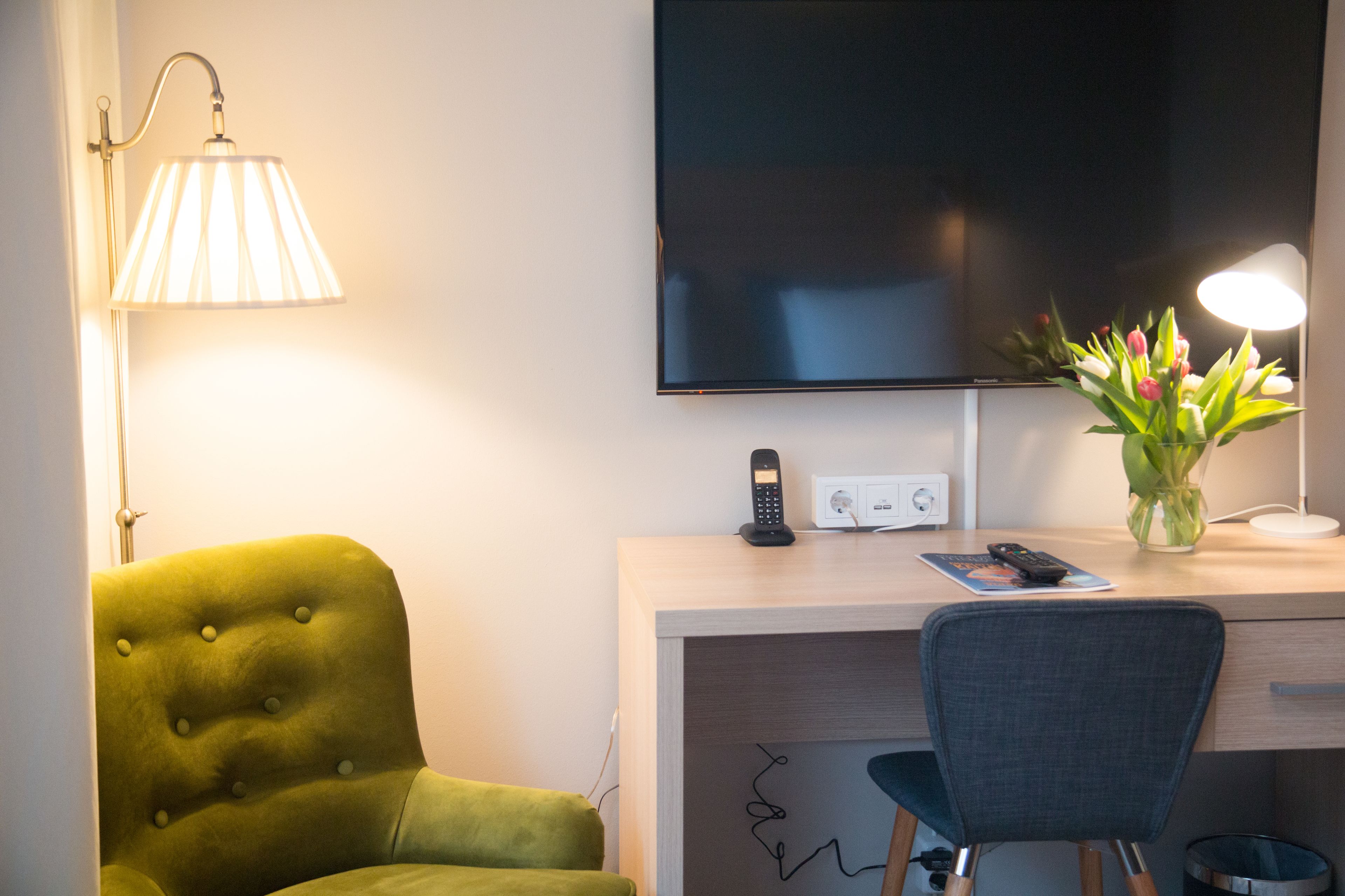 A hotel room with a green armchair, a desk with a lamp, a flat-screen TV, a vase with flowers, and a remote control on the desk.