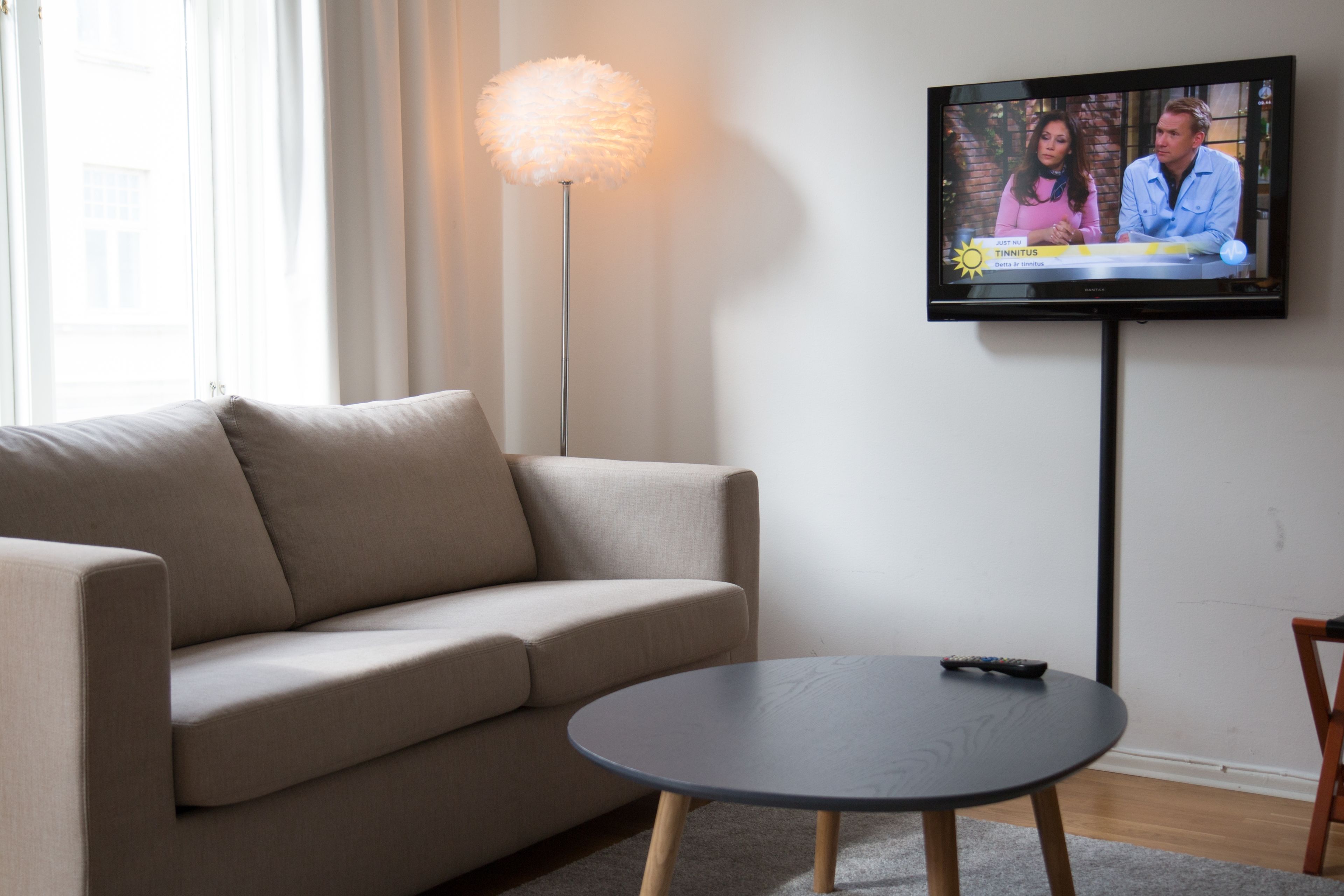 A living room with a beige sofa, a round coffee table, and a television mounted on the wall displaying a news program.