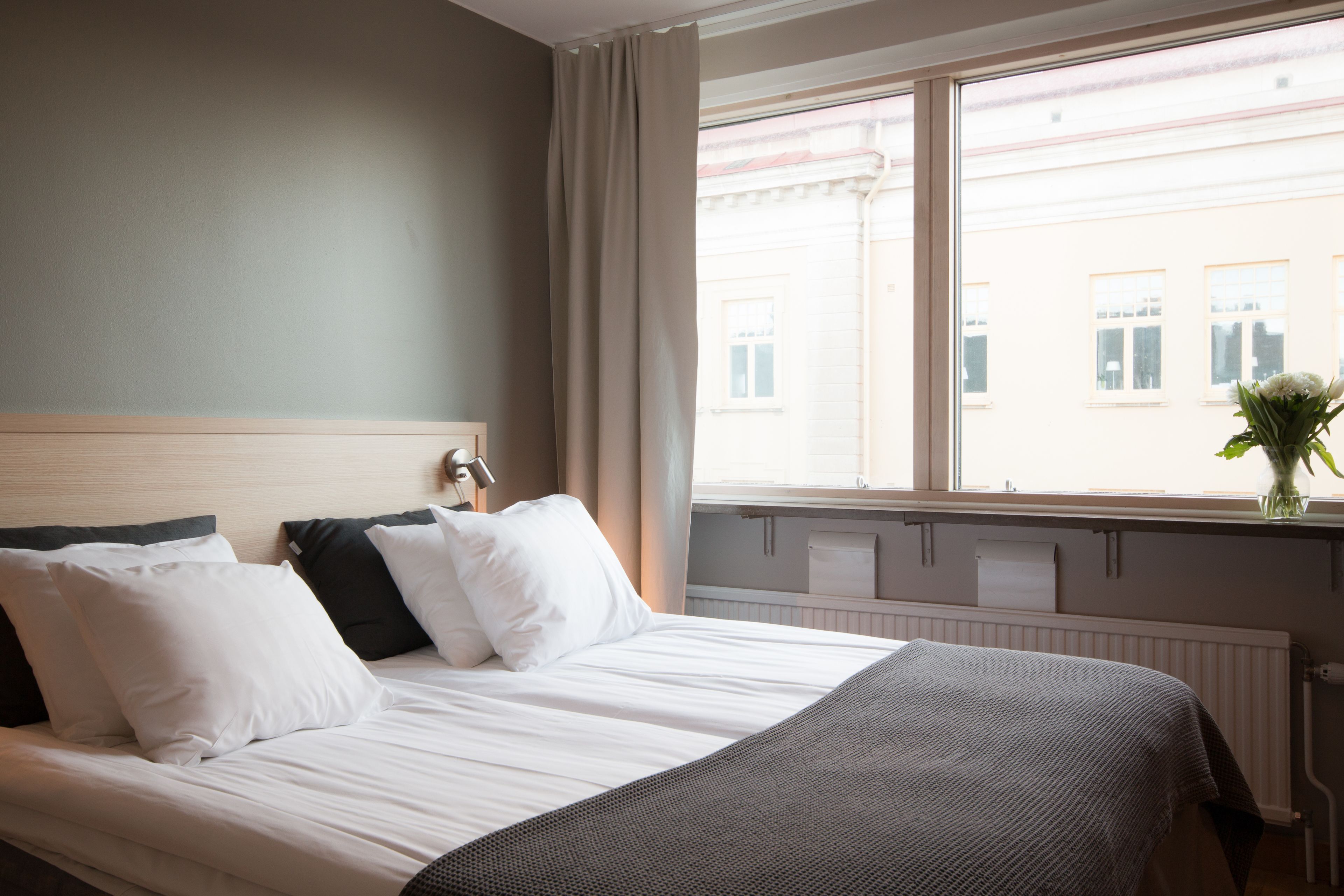 A hotel room with a bed, a window with curtains, a radiator, and a vase with flowers on the windowsill.