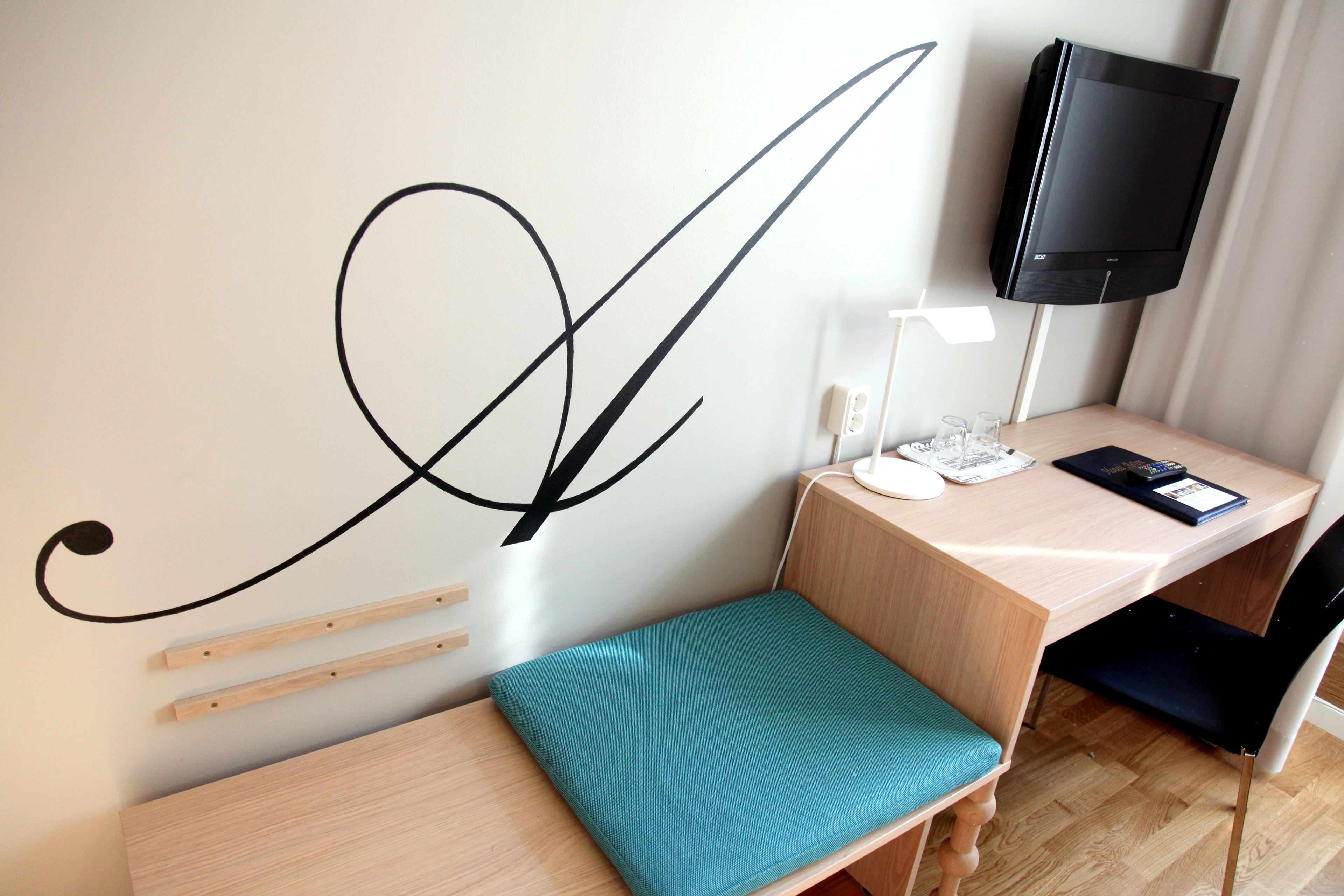 Une pièce de taille modeste, comprenant un bureau, un banc recouvert d’un tissu turquoise, un téléviseur fixé au mur et un mur orné d’un grand motif abstrait.