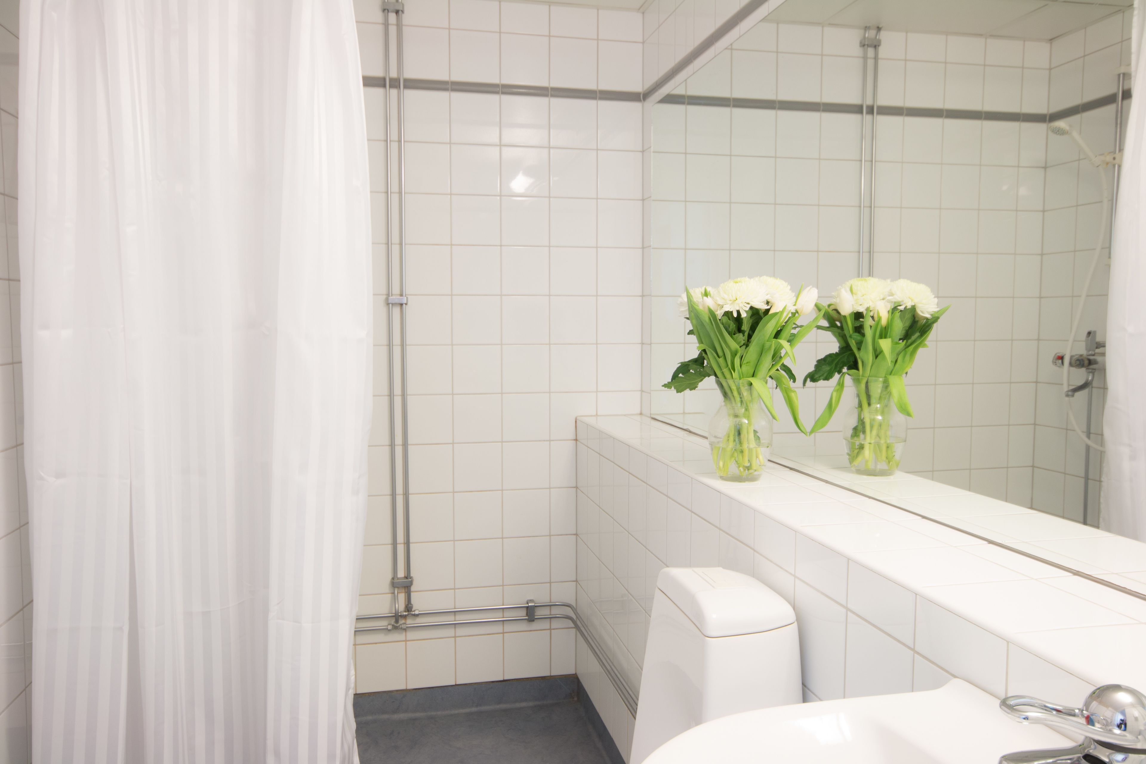 Un baño con azulejos blancos, una cortina de ducha, un inodoro y dos jarrones con flores sobre el lavamanos.