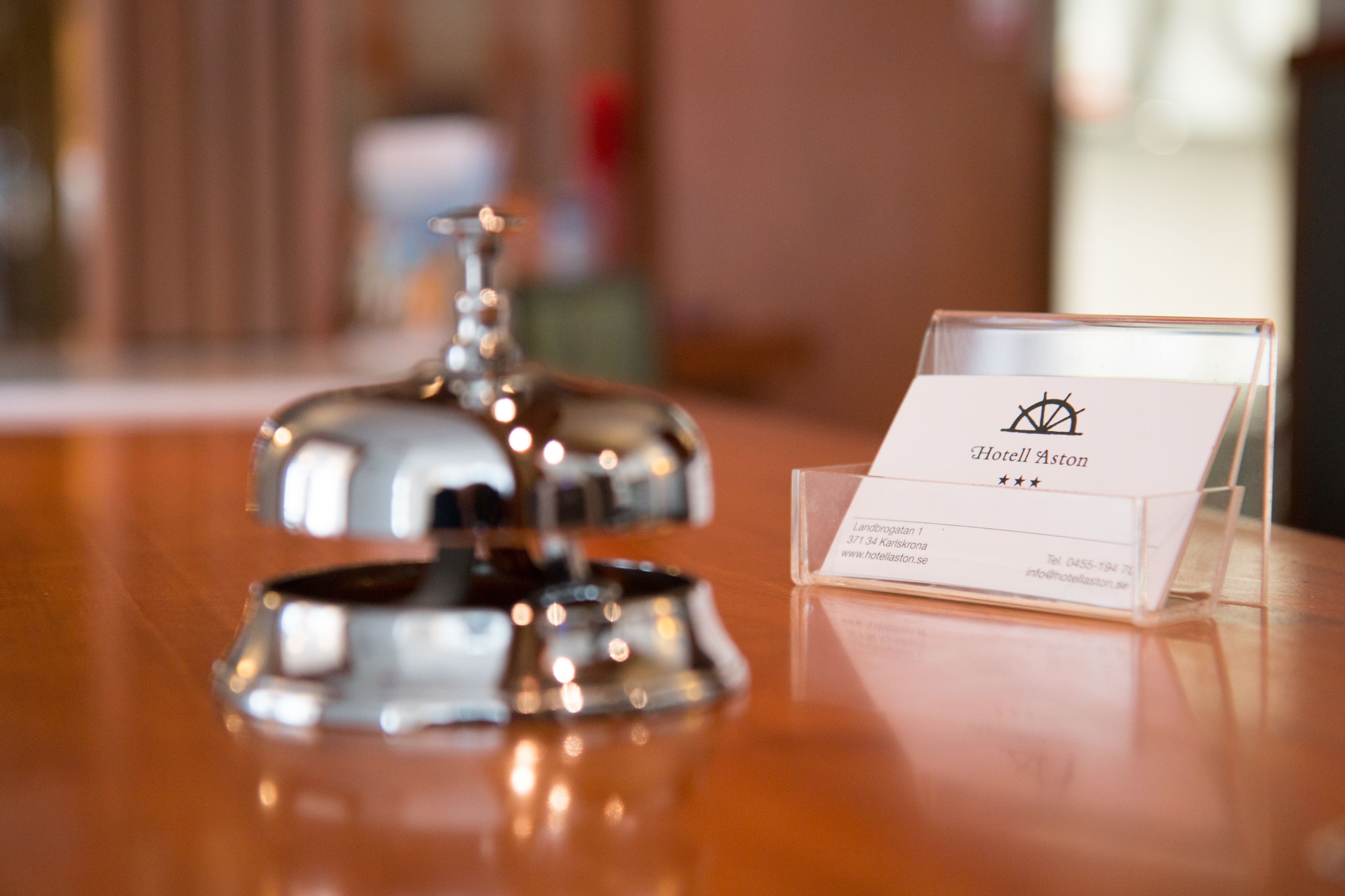 A hotel reception area with a polished wooden counter, a silver bell, and a business card holder displaying the Hotel Aston logo.