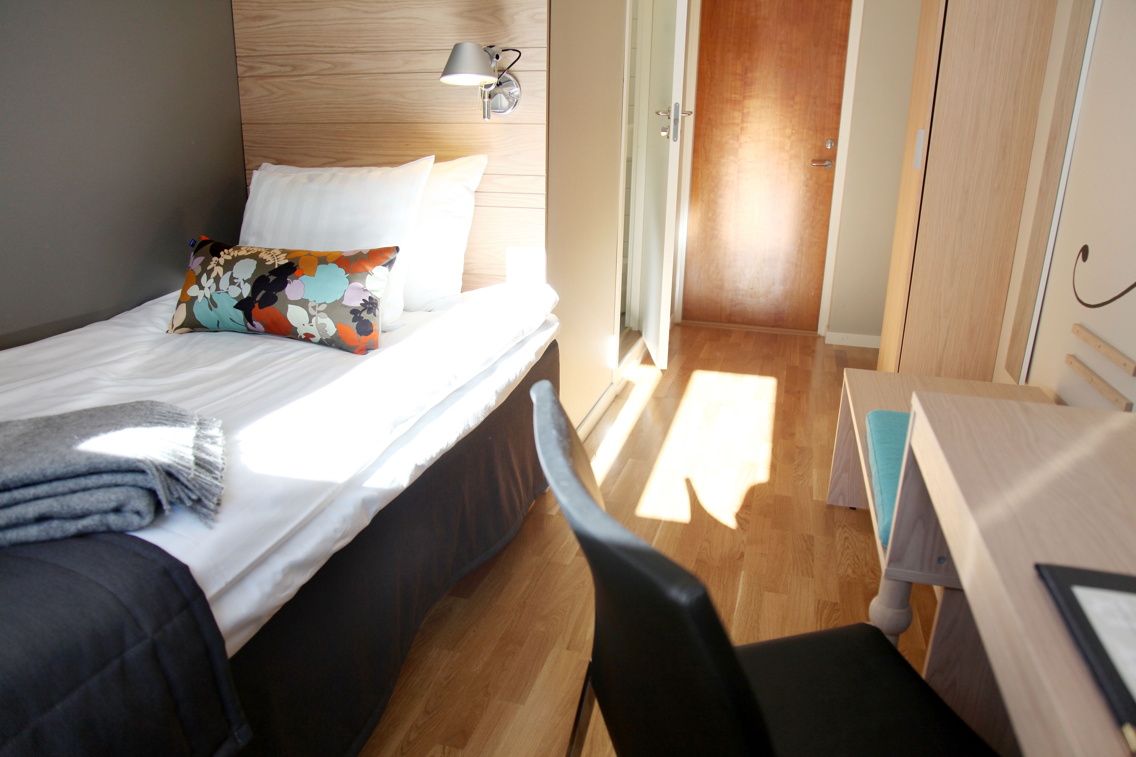 A hotel room with a bed, a desk, a chair, a lamp, and a wardrobe.