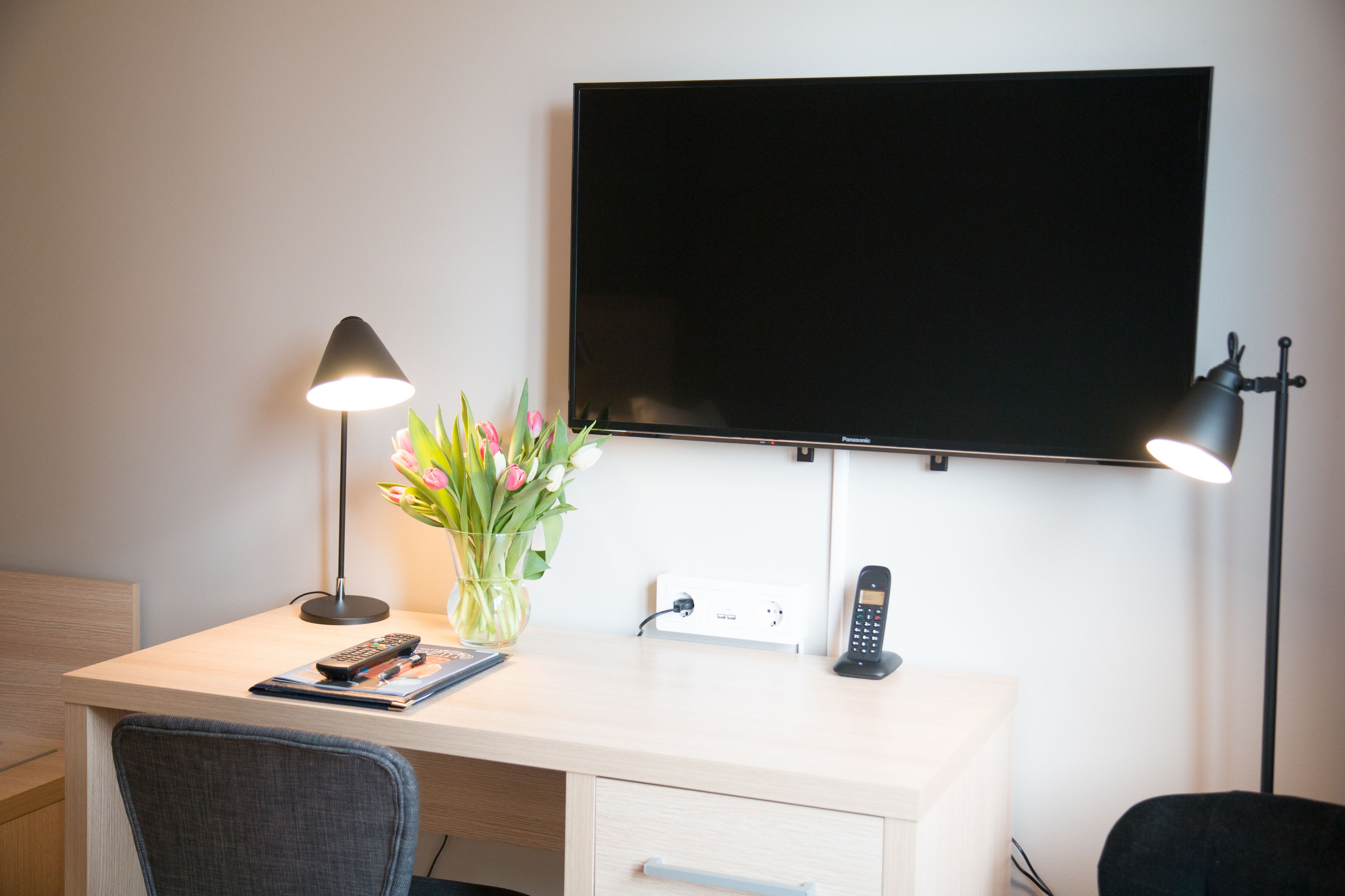 Habitació d'hotel amb escriptori, televisió de pantalla plana penjada a la paret, telèfon, làmpada i un ram de flors.