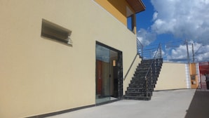Un edificio moderno con puerta de entrada de vidrio, una escalera con barandillas metálicas y cielo despejado con nubes.