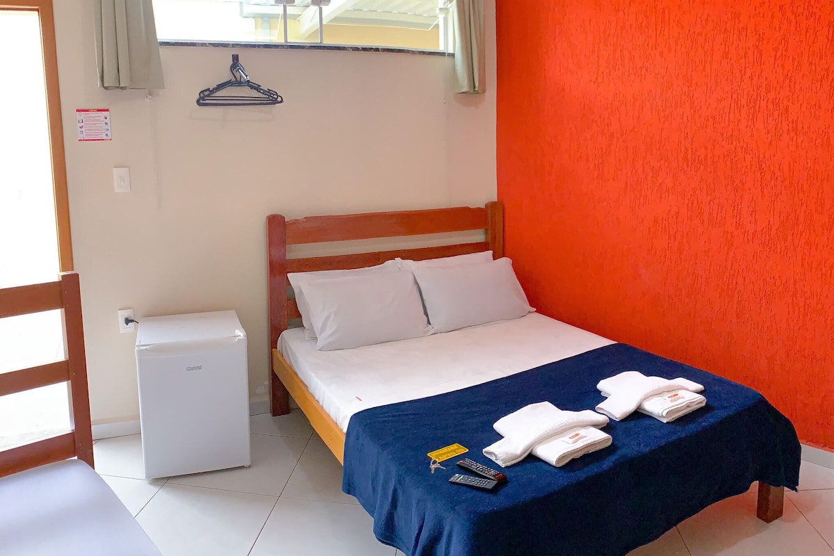 Quarto de hotel com cama, pequeno frigorífico branco e uma manta azul com toalhas e comando remoto.