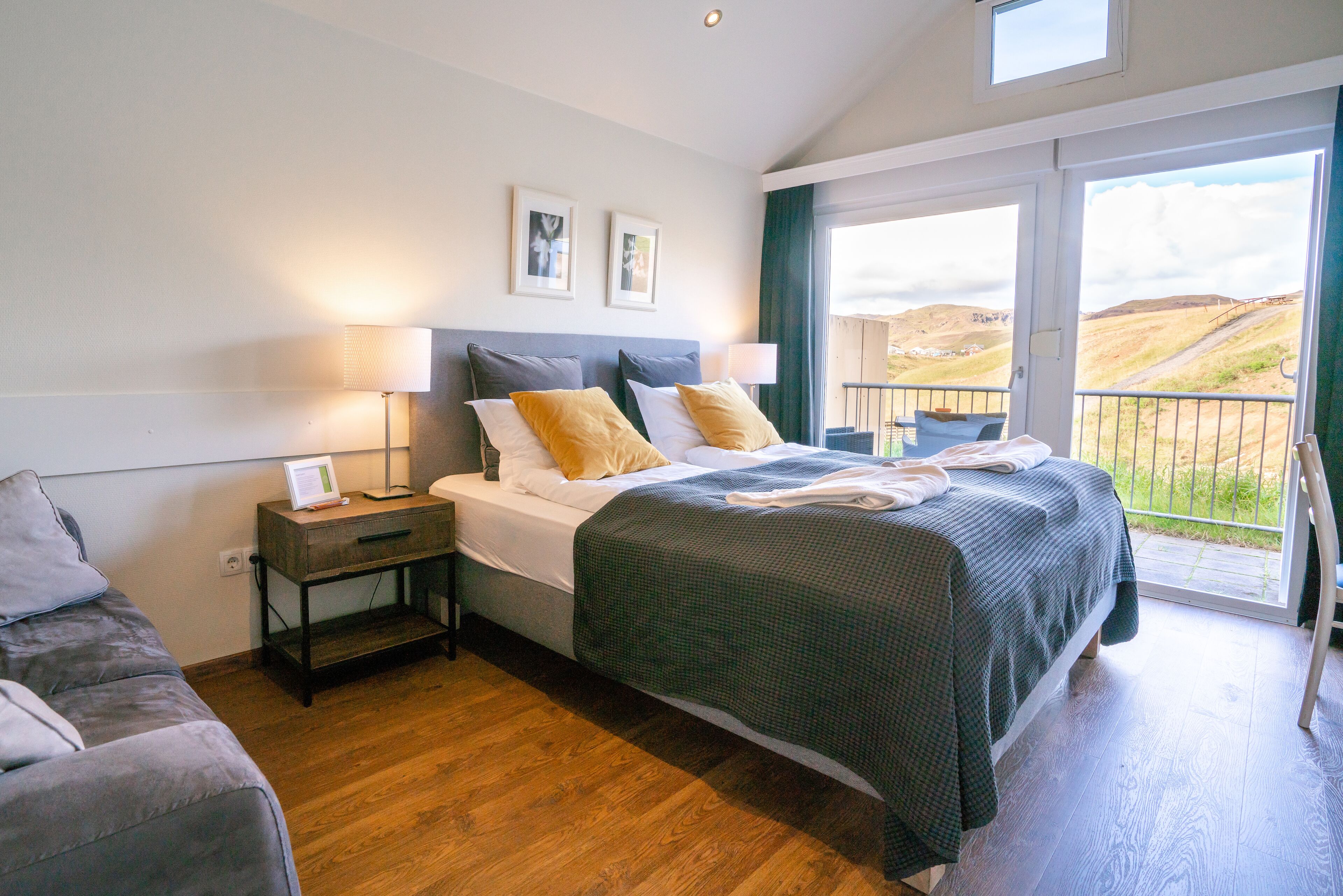 A bedroom with a bed, bedside tables, a nightstand, a sofa, and a balcony with a view of the countryside.