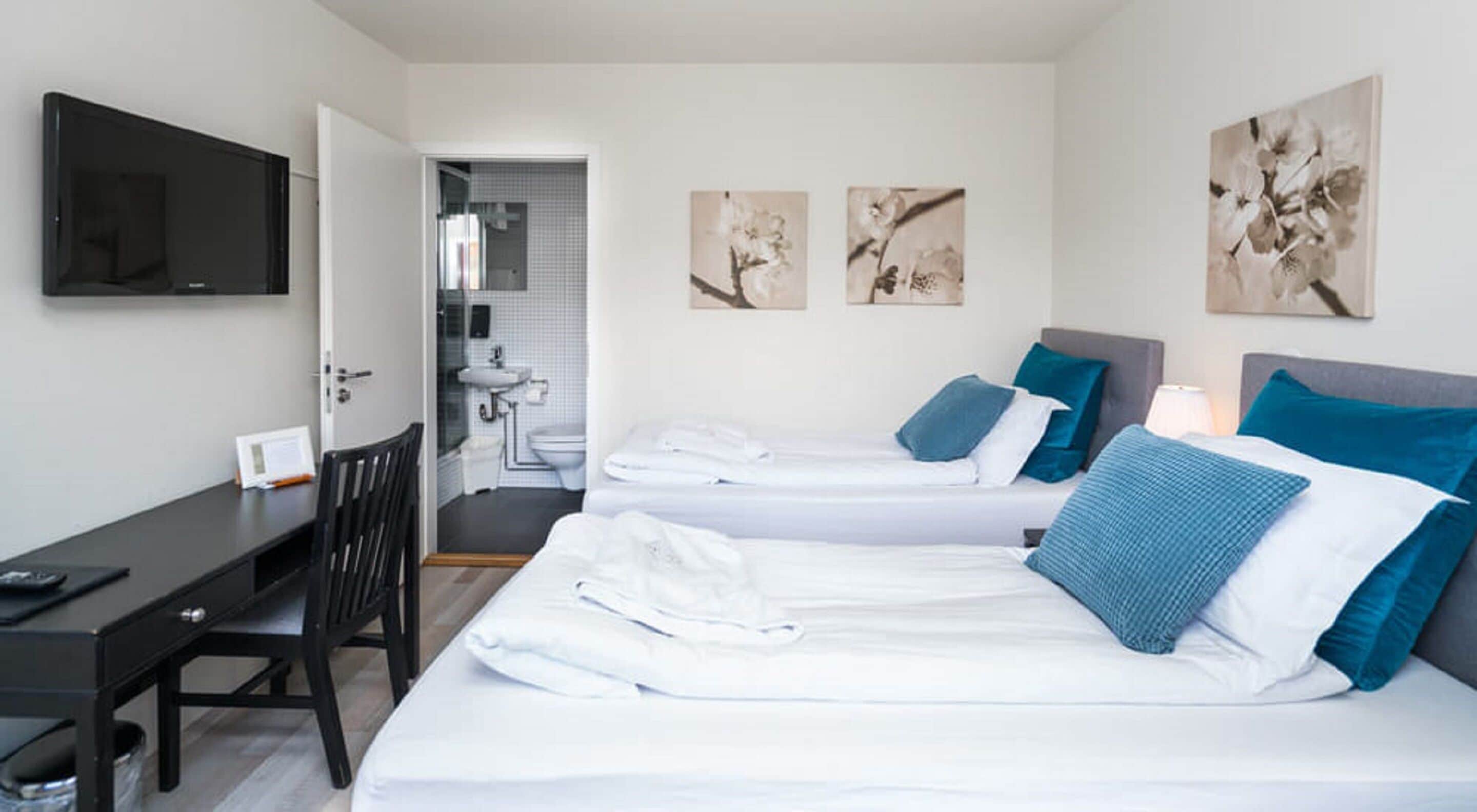 A hotel room with two beds, a desk, a chair, a TV, and artwork on the walls.