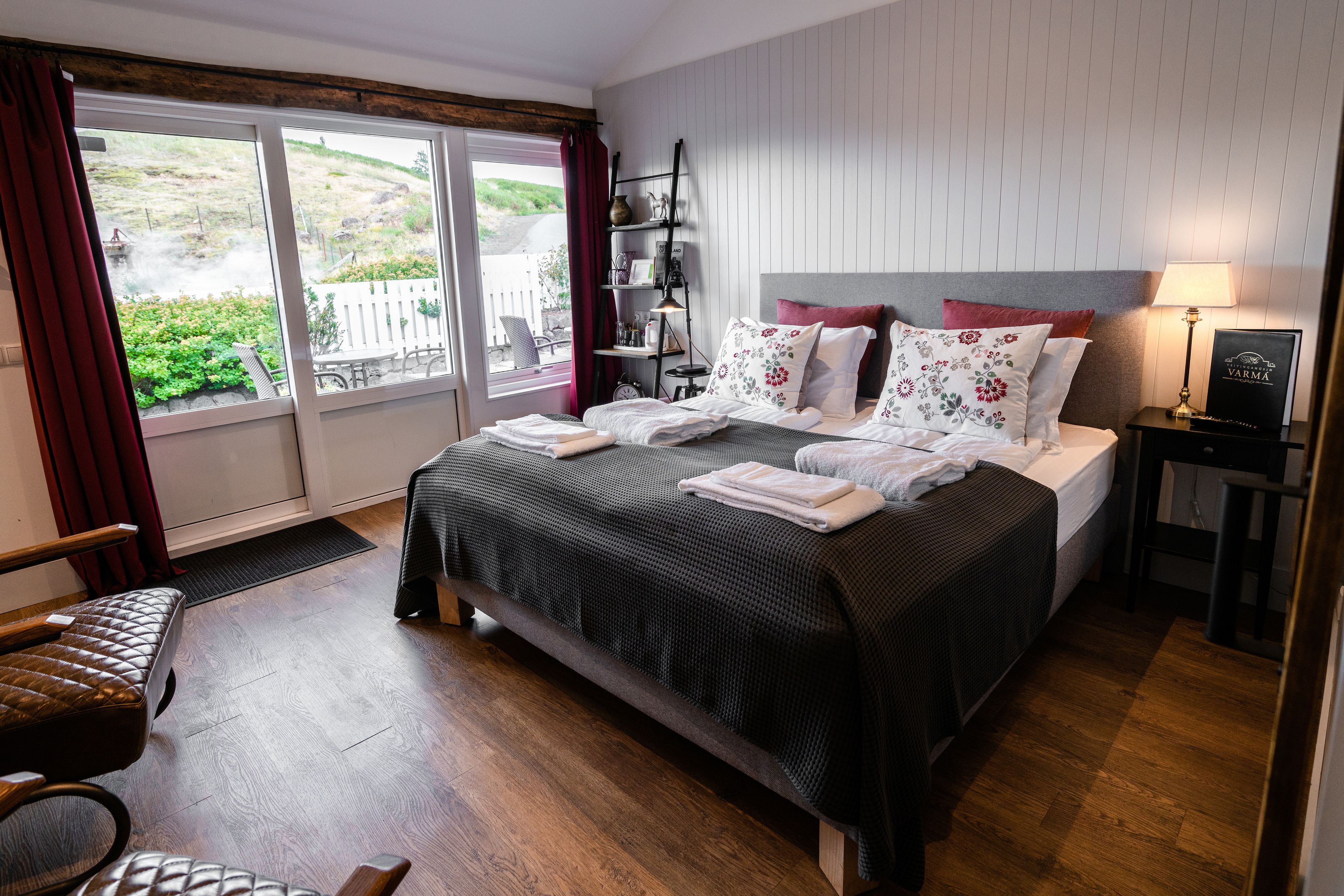 Cama bem arrumada com colcha escura e almofadas, mesa de cabeceira com abajur, cadeira de madeira e vista para um jardim pela janela.