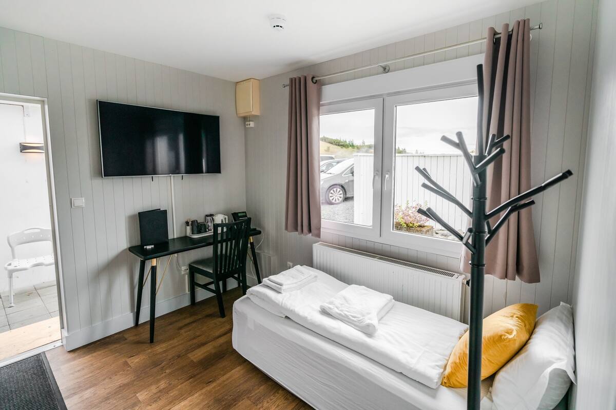 Una habitación pequeña con cama, escritorio, silla, televisor y ventana con cortinas.