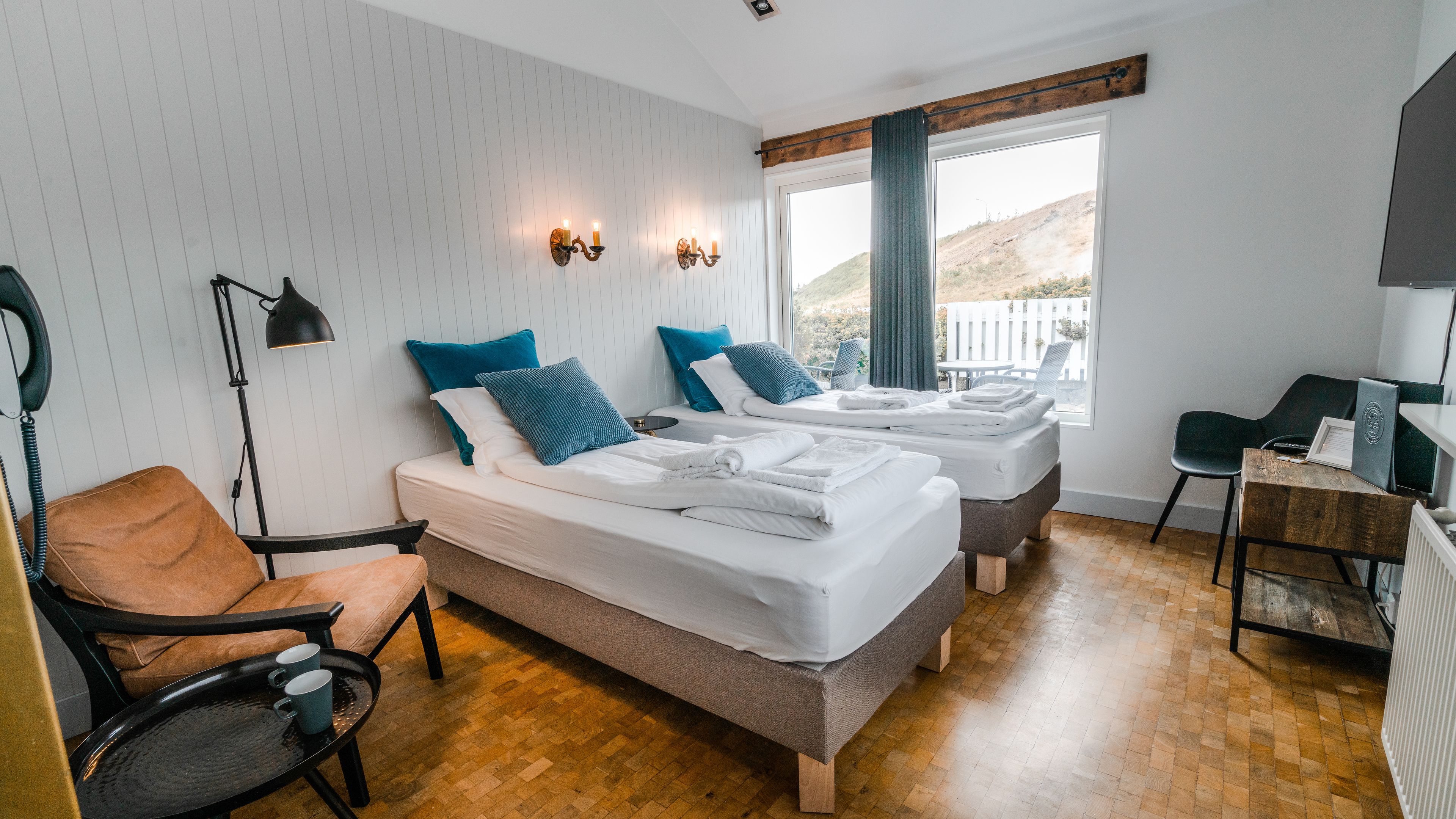 A hotel room with two beds, a desk, a chair, a lamp, and a window with a view of a white fence and hills.