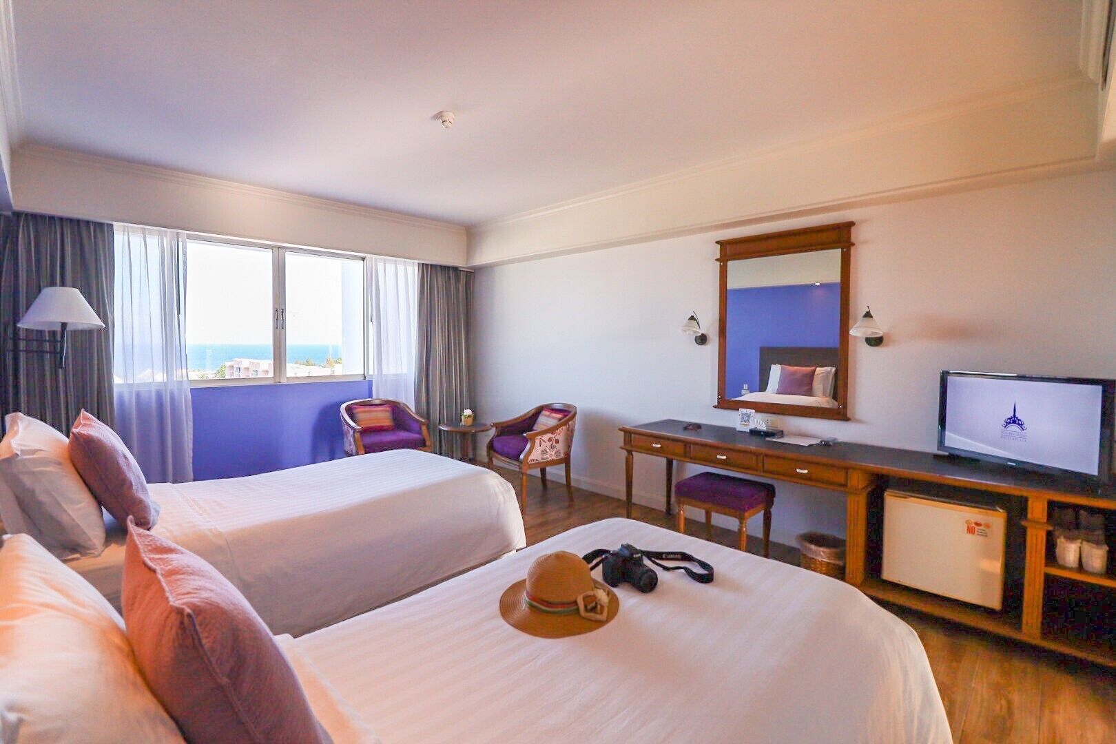 A hotel room with two beds, a desk with a computer, a chair, a small table, and a view of the sea.