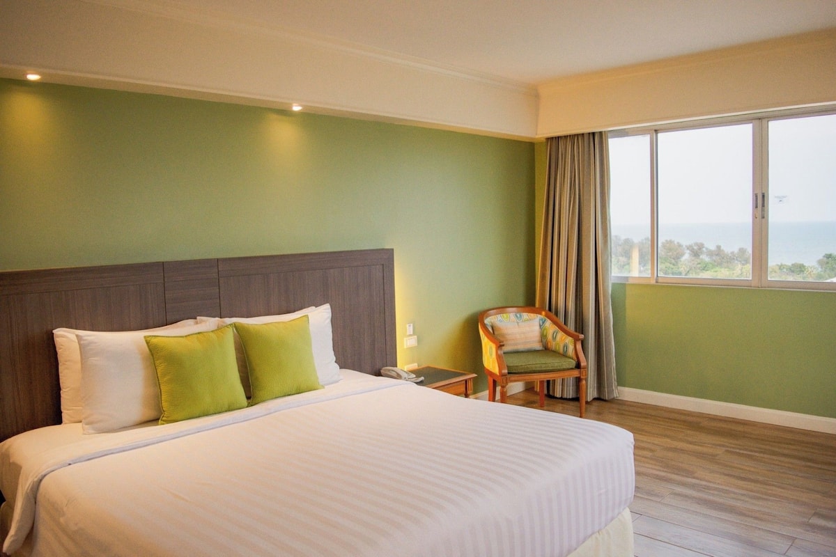 A hotel room with a large bed, a chair, a nightstand, and a view of the sea.