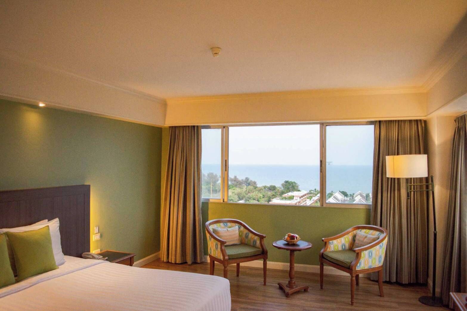 A hotel room with a bed, two chairs, a small table, a lamp, and a view of the sea.