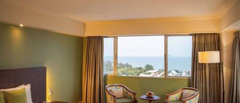 A hotel room with a bed, two chairs, a small table, a lamp, and a view of the sea.