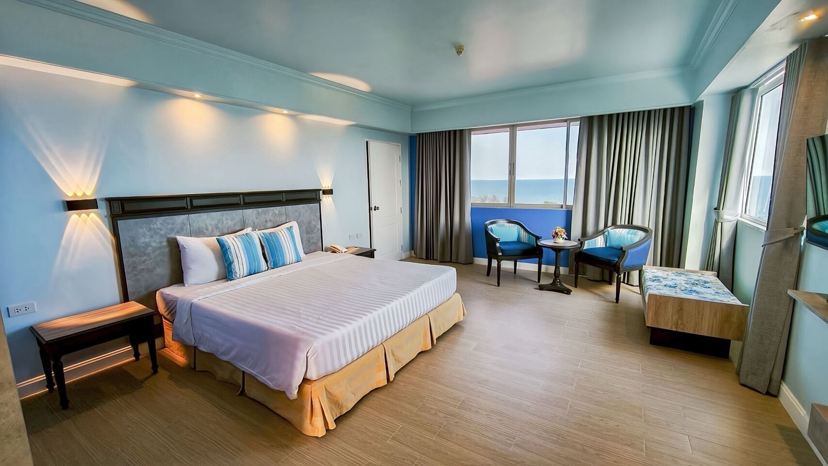 A hotel room with a large bed, a bedside table, a chair, a small table with a vase, and a view of the sea through the window.