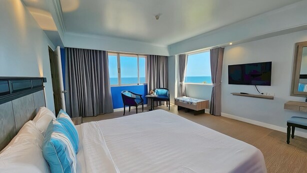 A hotel room with a large bed, a desk, a chair, a TV, and a view of the sea. - HUA HIN GRAND HOTEL AND PLAZA (Hua Hin)
