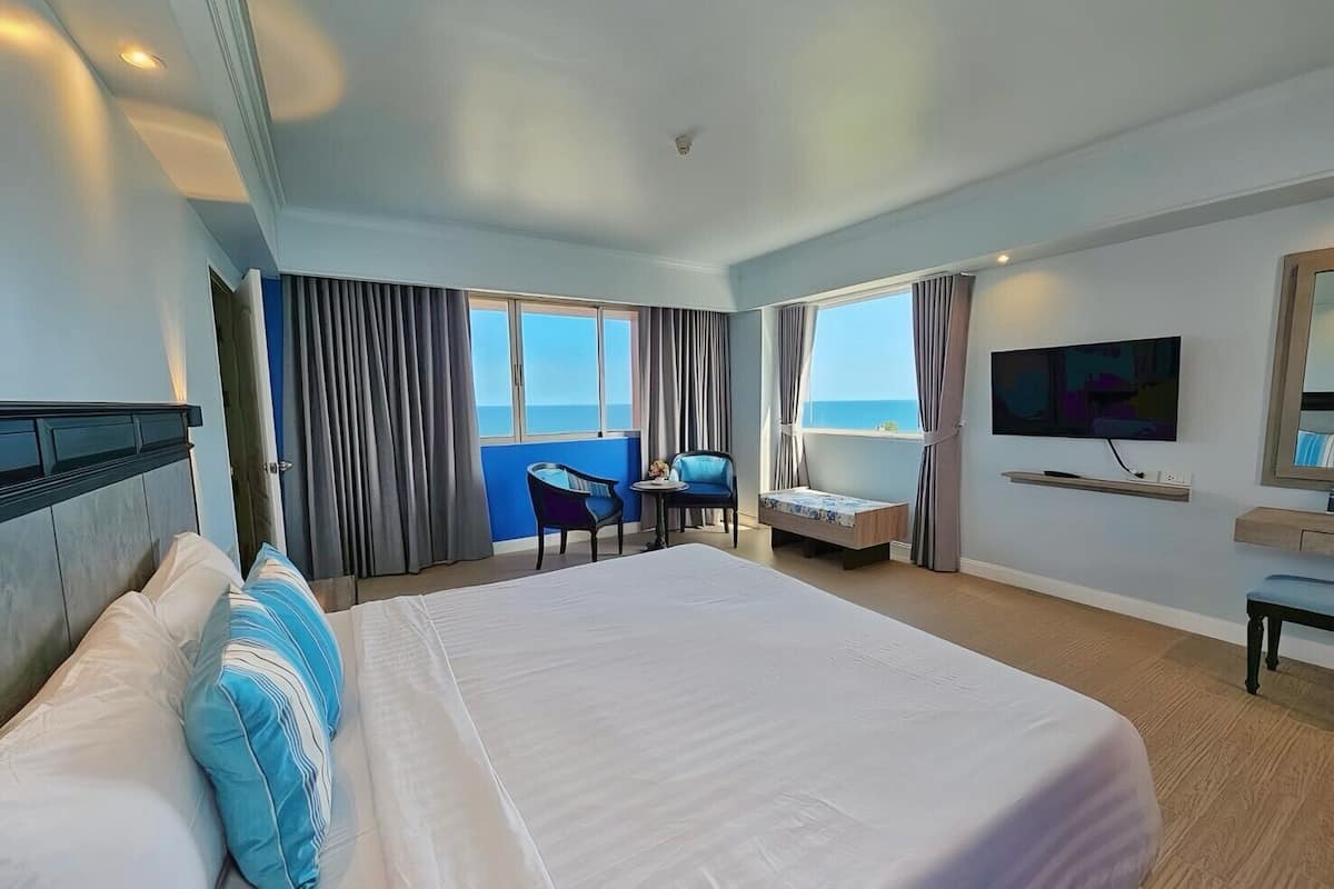 A hotel room with a large bed, a desk, a chair, a TV, and a view of the sea.