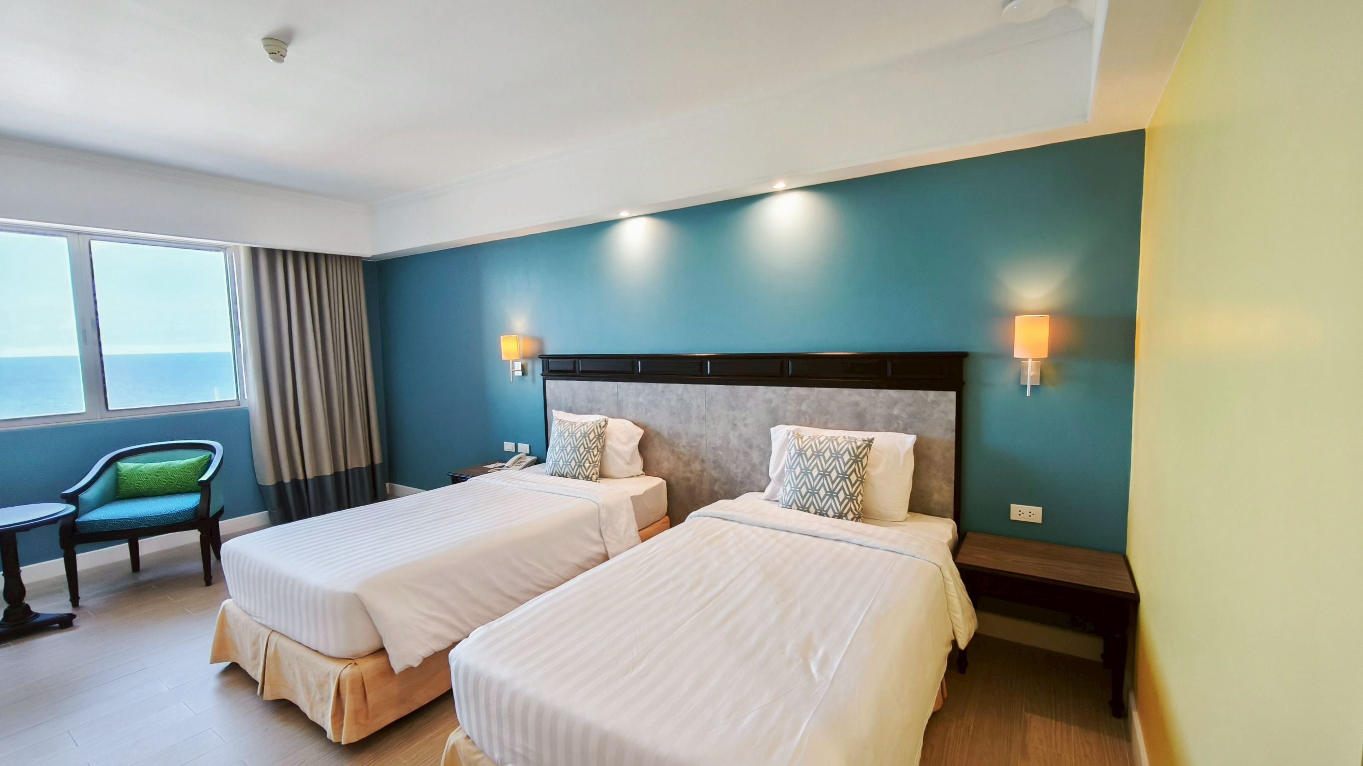 A hotel room with two beds, a chair, a table, and a view of the sea.