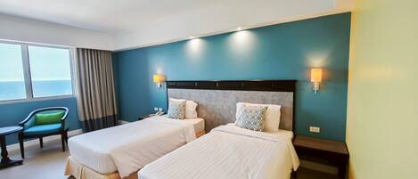 A hotel room with two beds, a chair, a table, and a view of the sea.