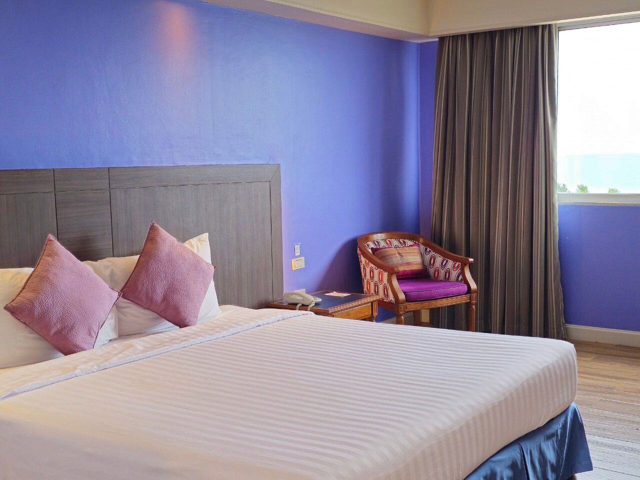 A hotel room with a large bed, a wooden headboard, a chair with a patterned cushion, and a view of the outdoors through a window with curtains.