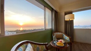 A hotel room with a view of a sunset over a body of water, a sofa, a chair, a small table with a banana and a glass of water, and a lamp. - HUA HIN GRAND HOTEL AND PLAZA (Hua Hin)
