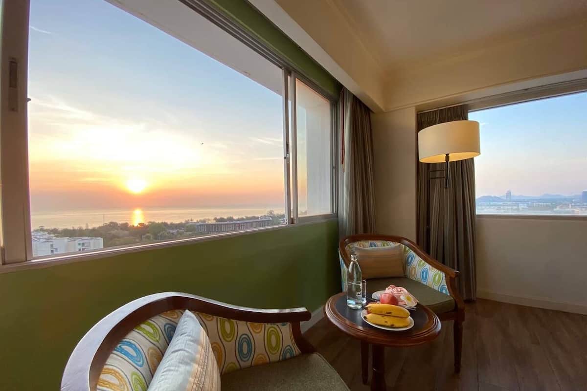 A hotel room with a view of a sunset over a body of water, a sofa, a chair, a small table with a banana and a glass of water, and a lamp.