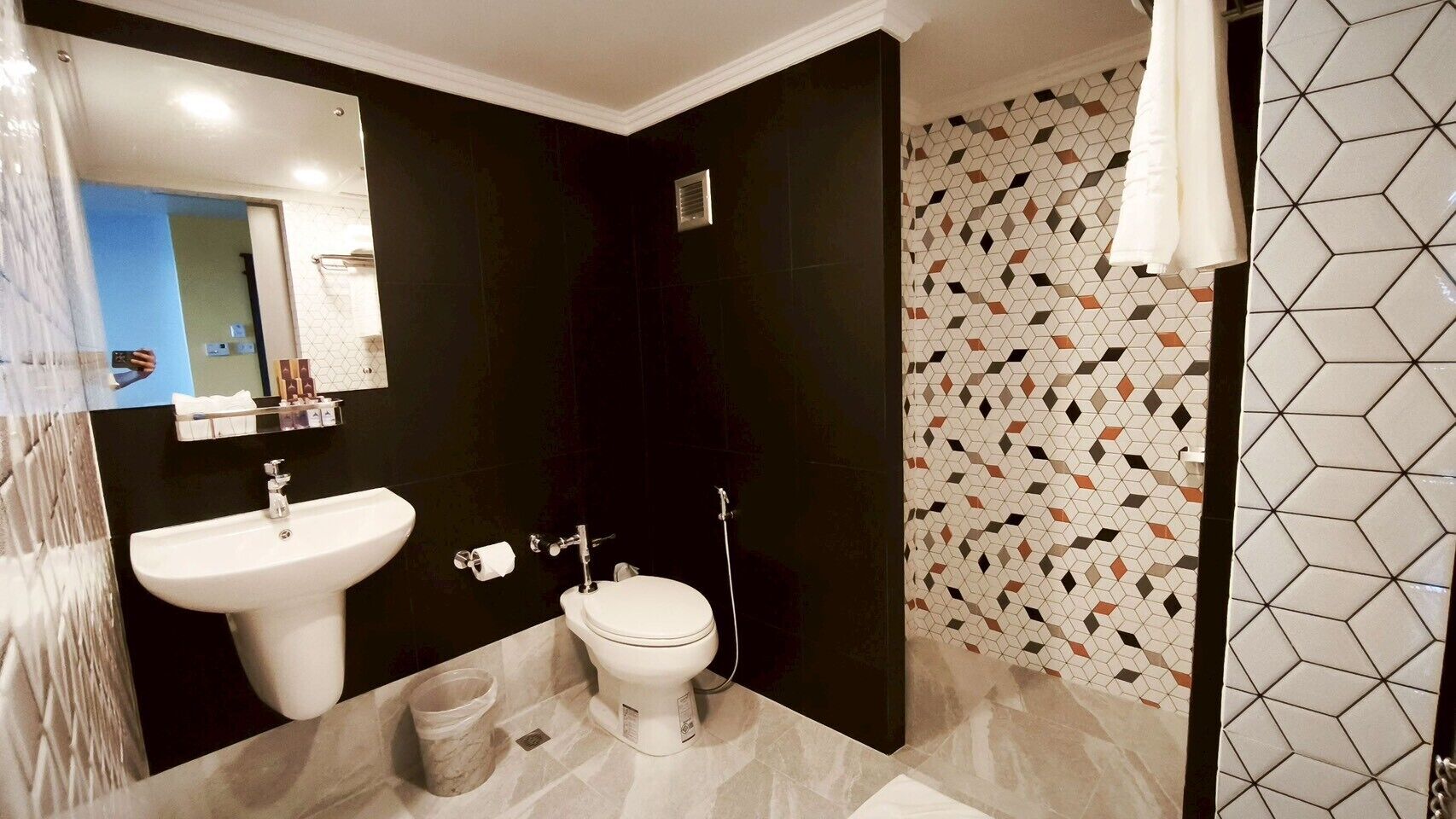 A modern bathroom with a white sink, toilet, and shower. The walls are dark with geometric patterned tiles.