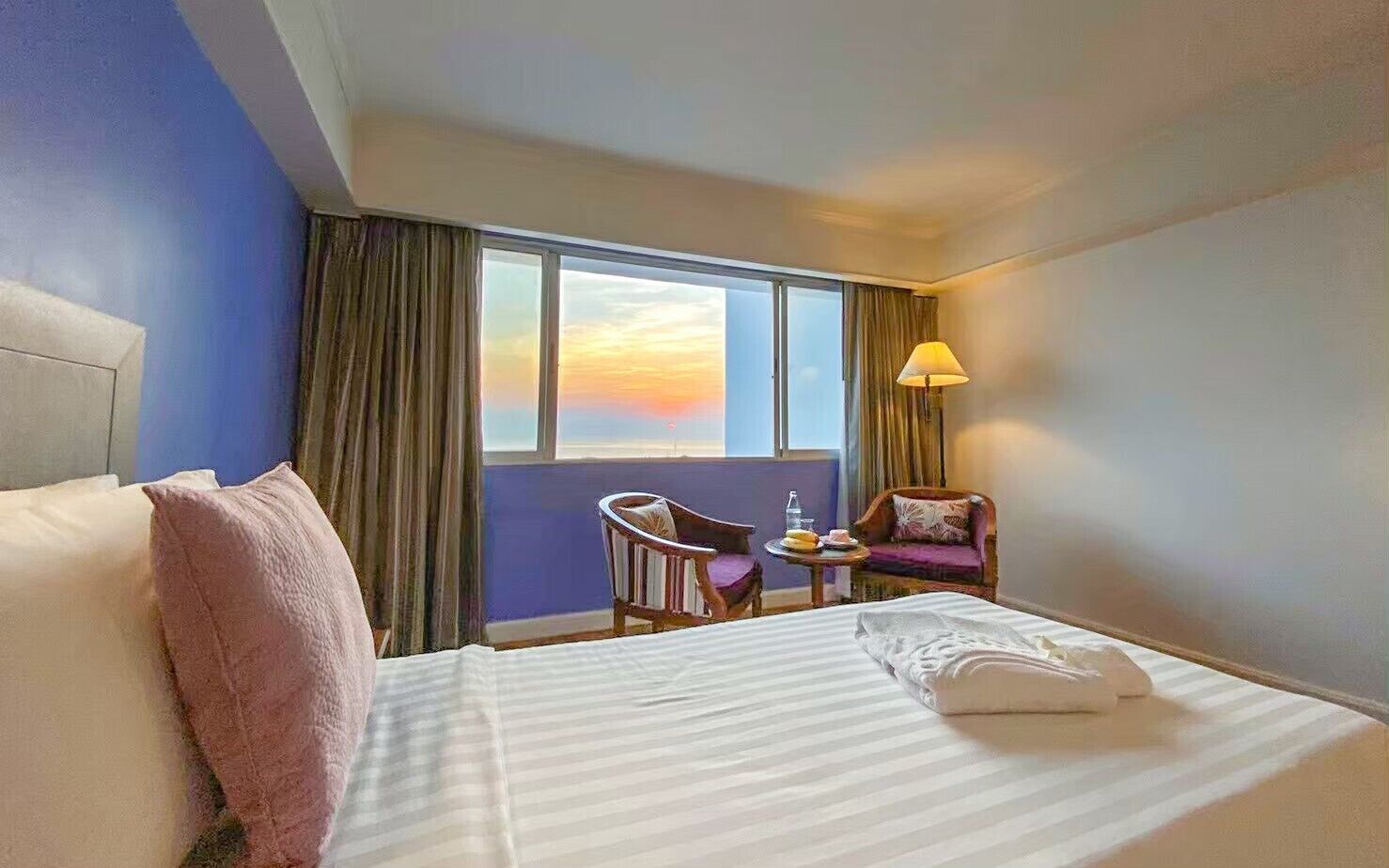 A hotel room with a large bed, a chair, a small table with a lamp, and a view of the sunset through the window.