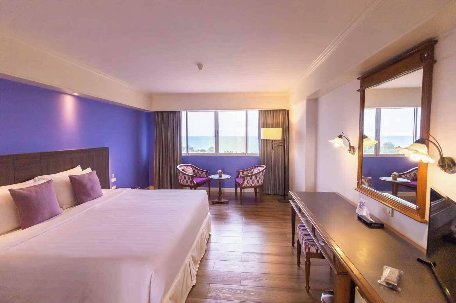 A hotel room with a bed, two chairs, a desk, a mirror, and a view of the sea.