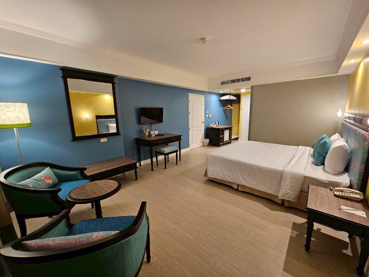 A hotel room with a bed, two chairs, a small table, a desk, a mirror, and a TV.