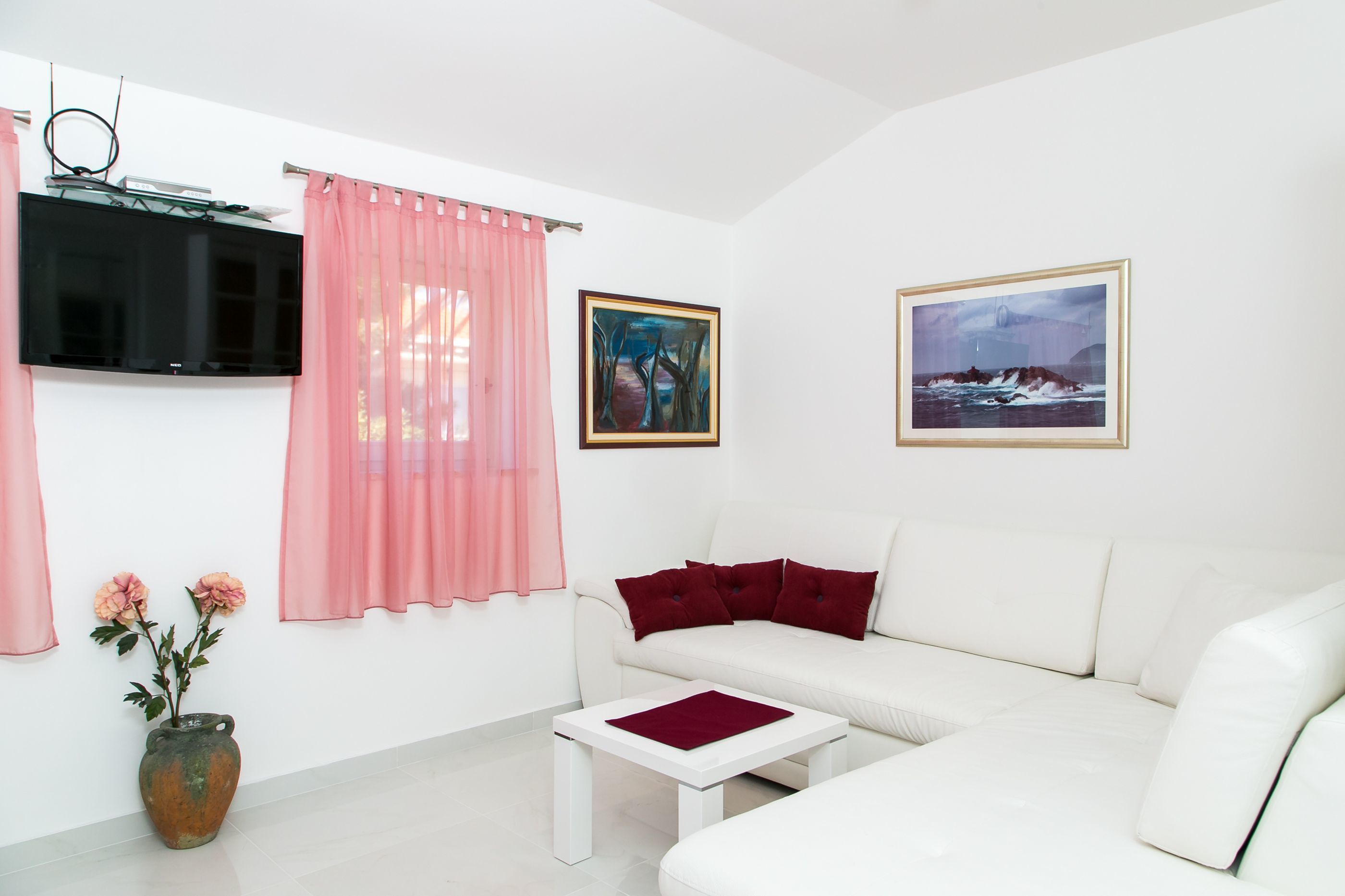 A modern living room with a white sofa, a flat-screen TV mounted on the wall, two framed artworks, a vase with flowers, and a pink curtain.