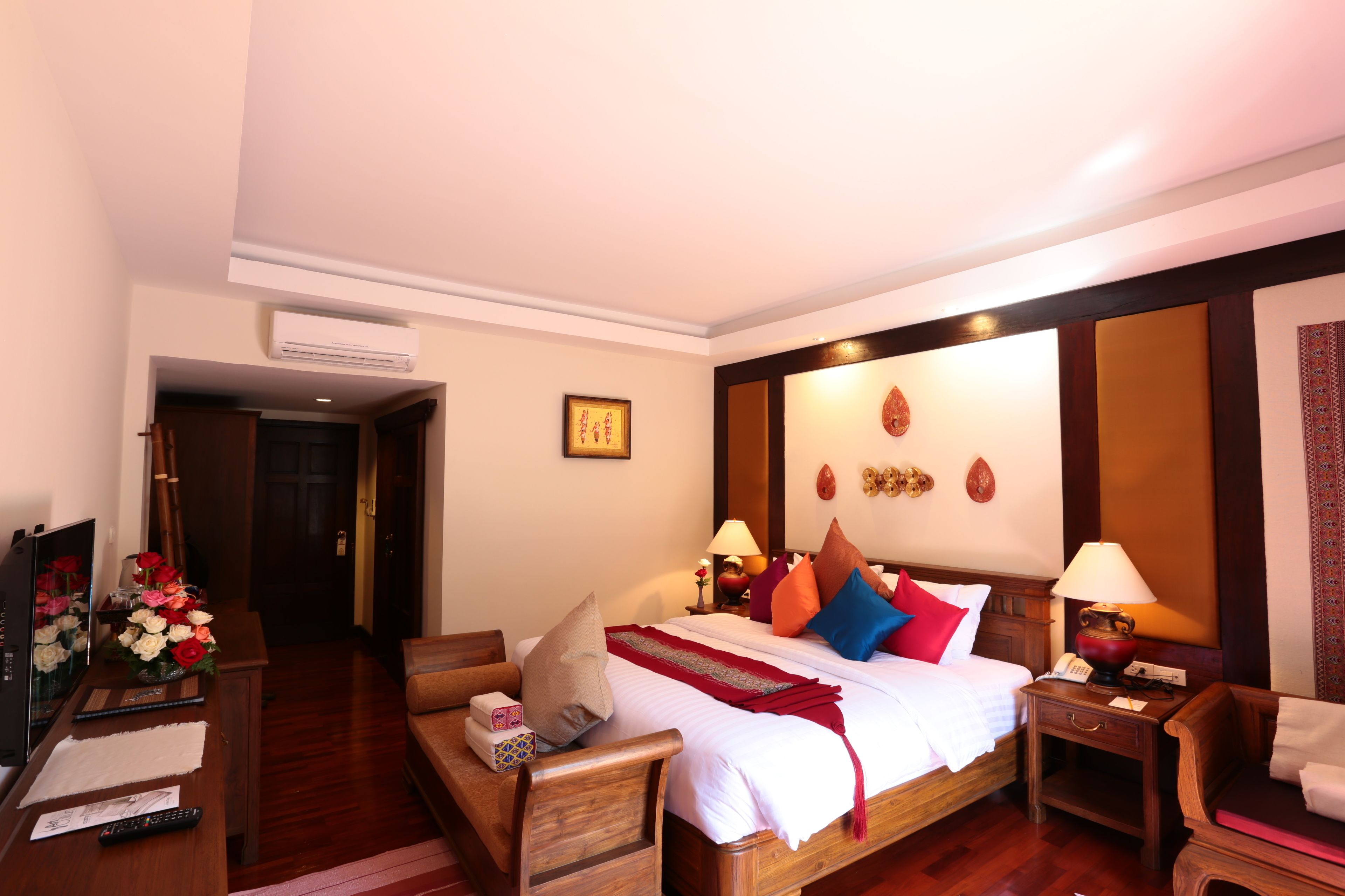Quarto de hotel com cama de madeira, mesas de cabeceira, escrivaninha com computador e controles remotos, e um espelho.