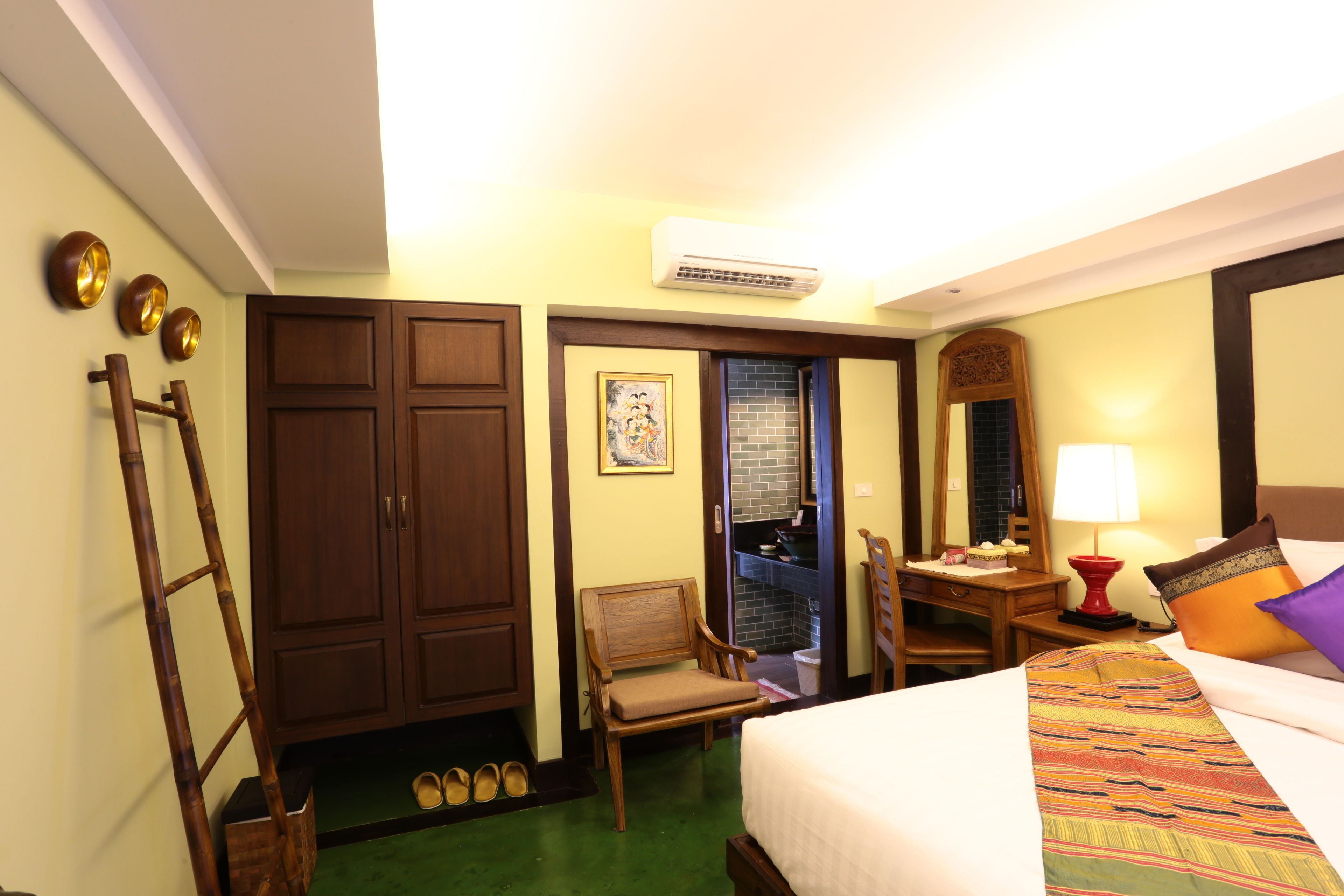 A hotel room with a bed, a desk with a lamp, a chair, a wardrobe, and an air conditioning unit.