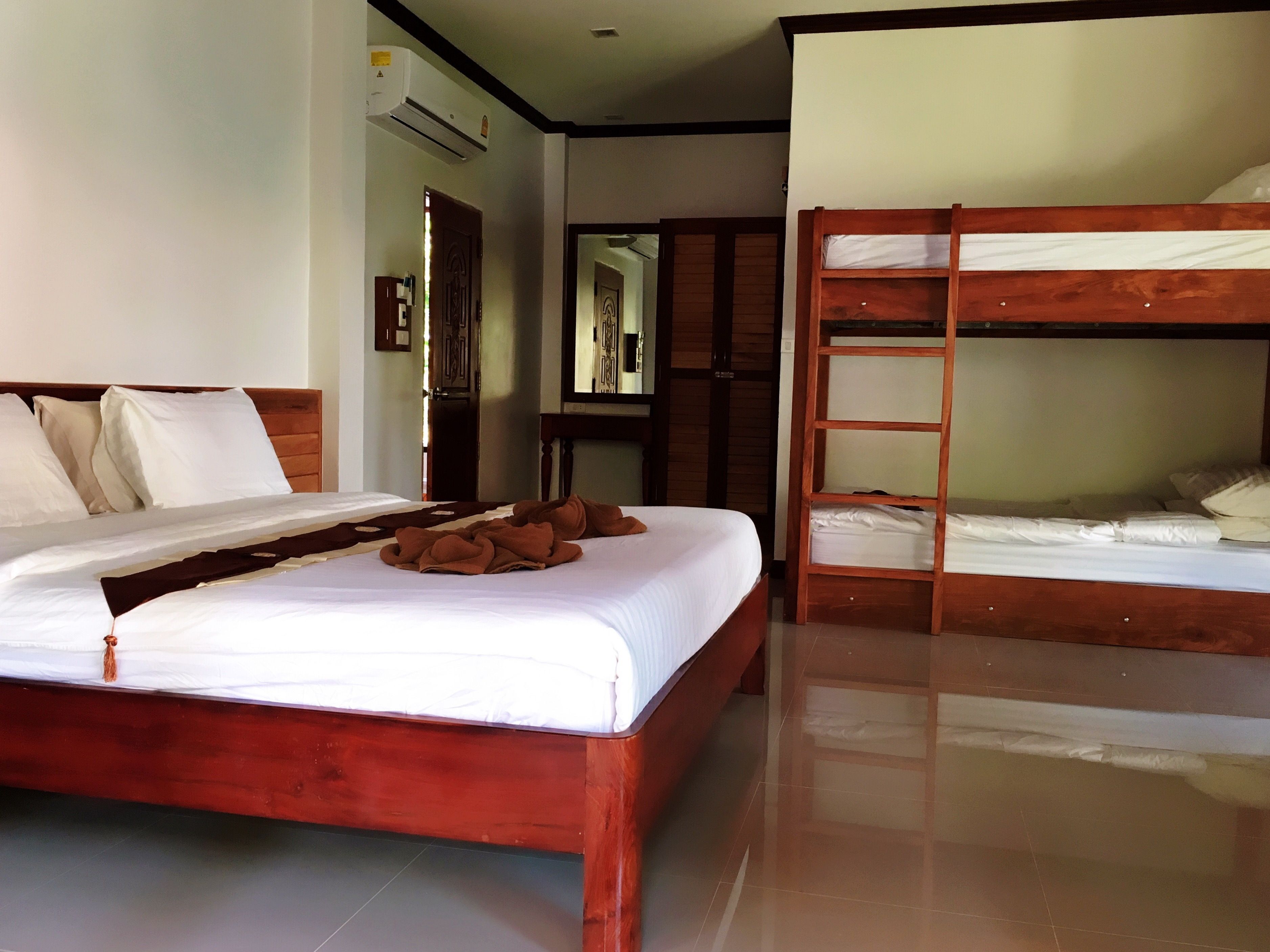A hotel room with a wooden bed, a bunk bed, and a bathroom with a mirror and towel rack.