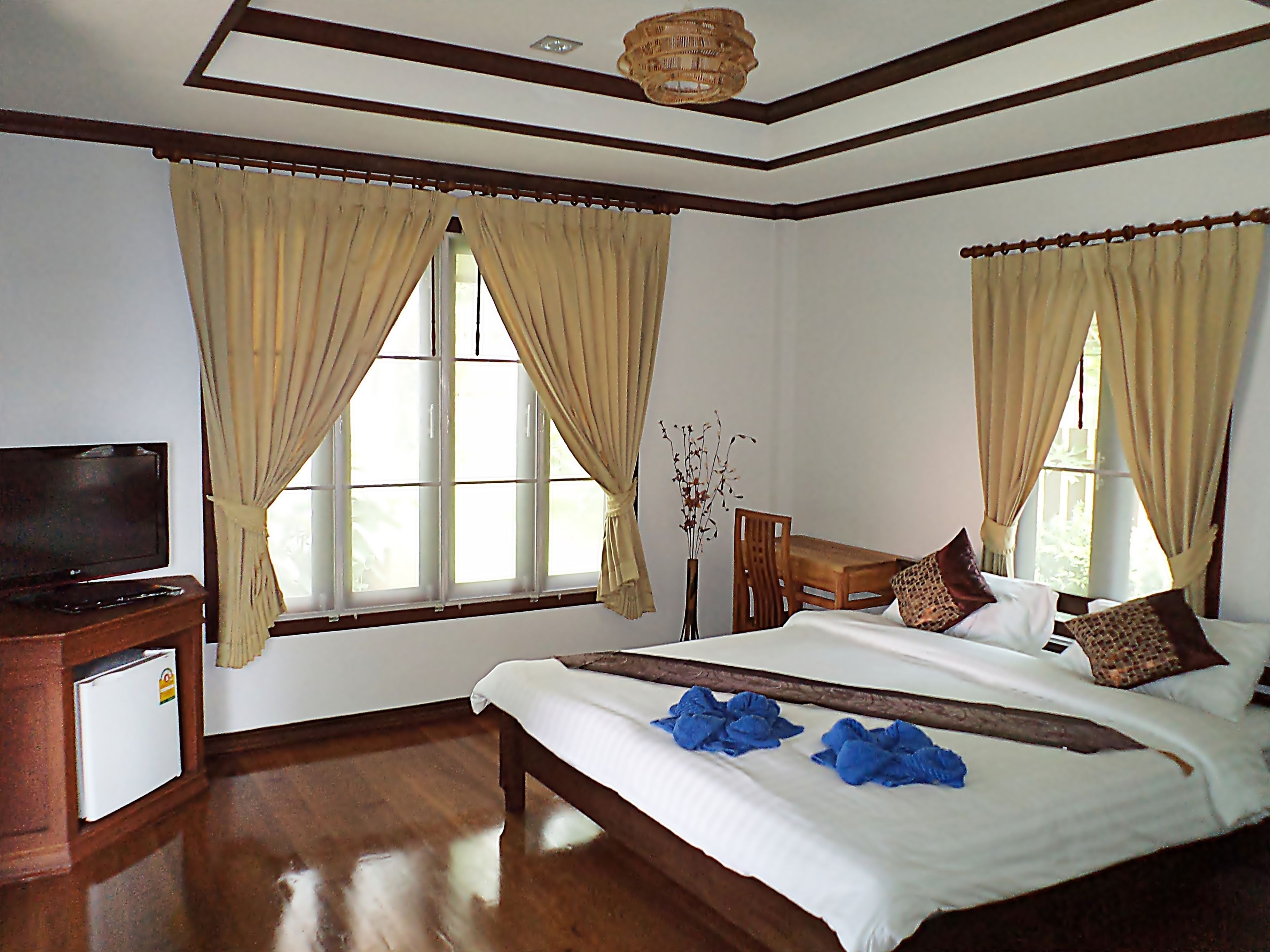 A hotel room with a bed, a desk, a television, and large windows with curtains.