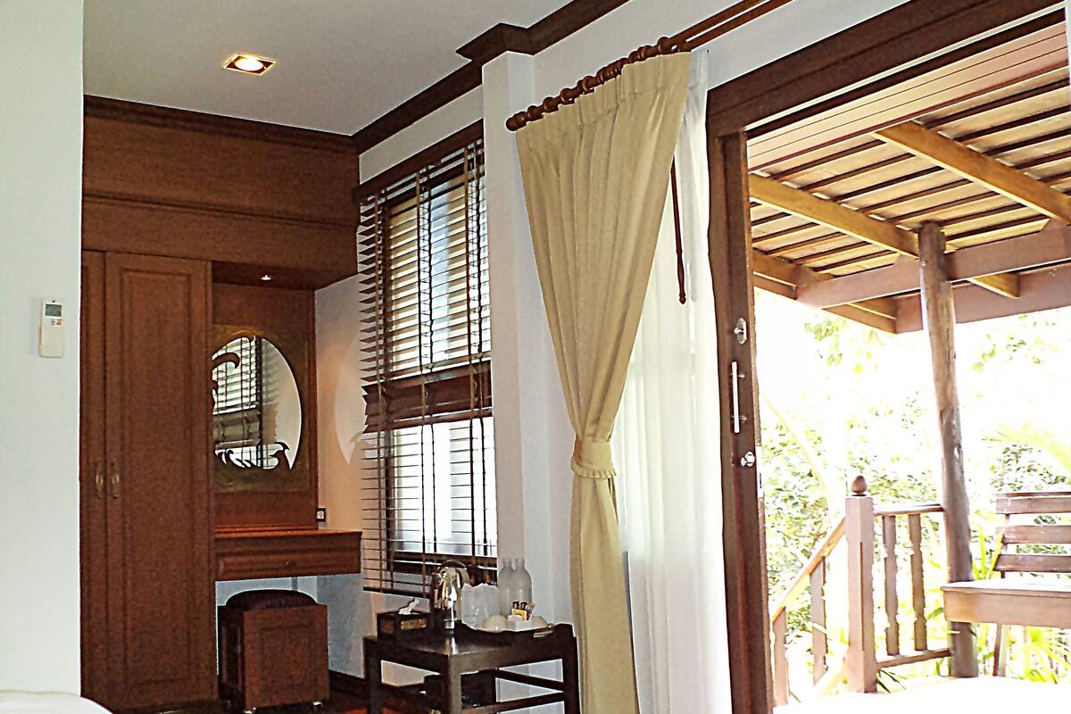 A room with a bed, a small table with a bottle and glass, a mirror, and a window with blinds.