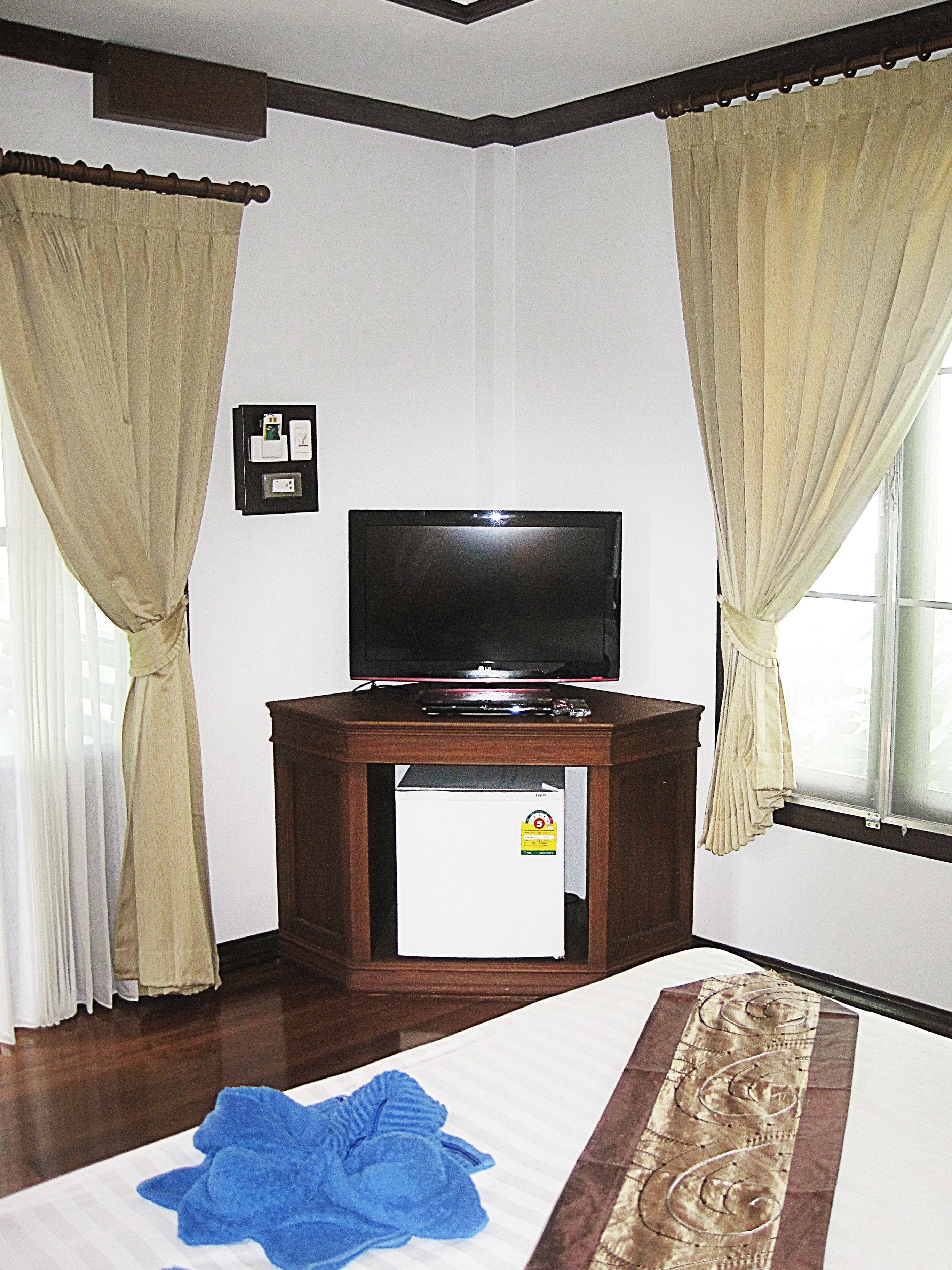 A hotel room with a flat-screen TV on a wooden stand, beige curtains, a bed with a patterned bedspread, and a small refrigerator.
