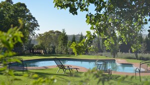 A swimming pool with a grassy area, a wooden deck, and a metal chair. - Valverde Country Hotel (Muldersdrift)