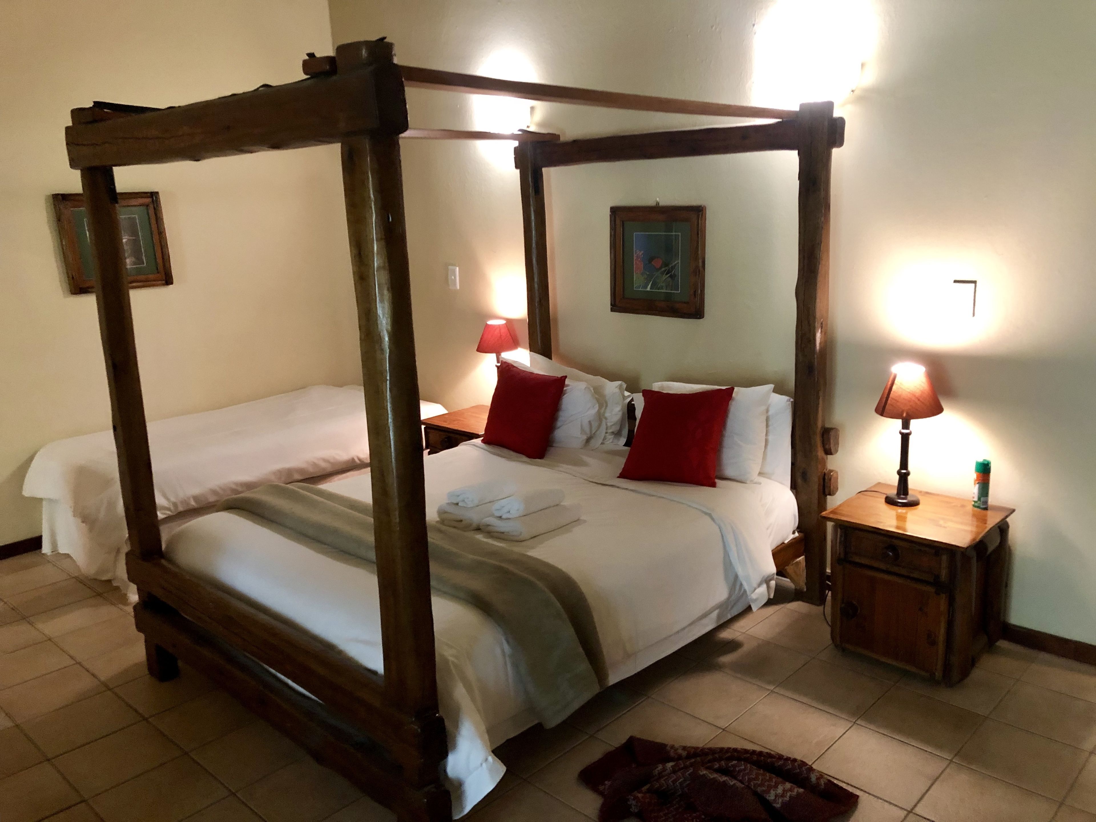 Un lit à baldaquin avec deux oreillers et deux coussins décoratifs rouges, une table de chevet en bois avec une lampe, et un tableau encadré accroché au mur.