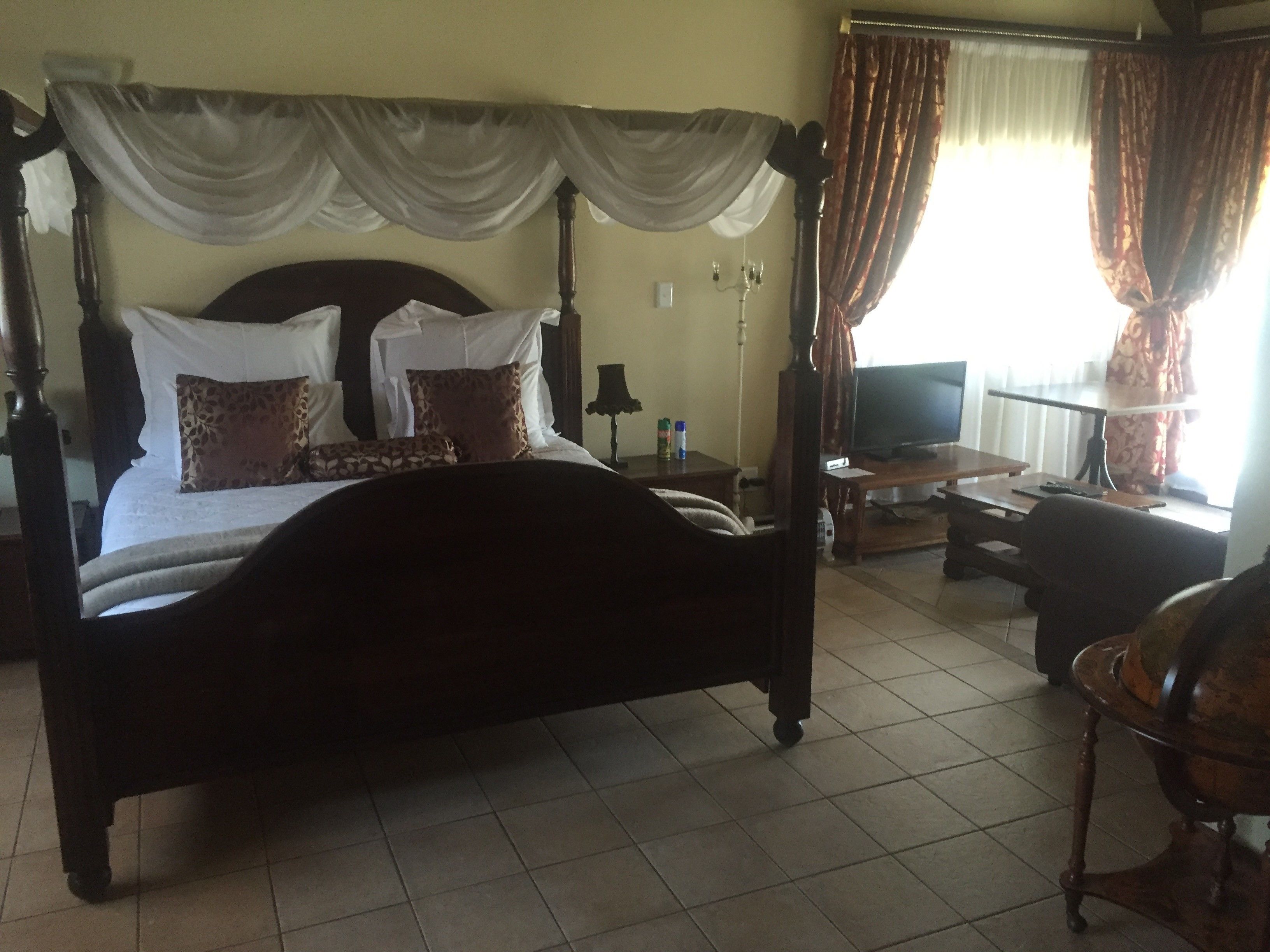 A four-poster bed with a canopy, a television, a desk, and a chair in a room with tiled flooring.